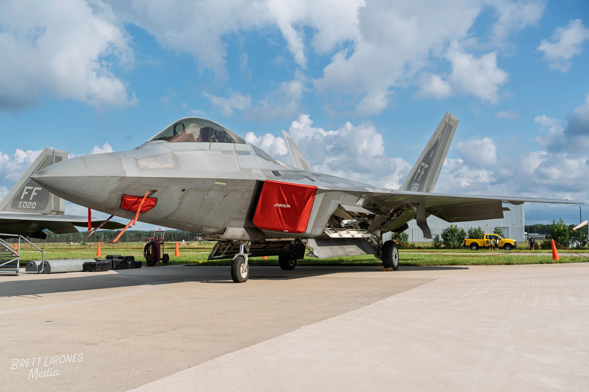 A military fighter jet parked outdoors on a tarmac with an open maintenance panel. The aircraft has a gray camouflage paint, a clear canopy, and a red cover over the intake. The background shows a partly cloudy sky, a grassy field, and vehicles.