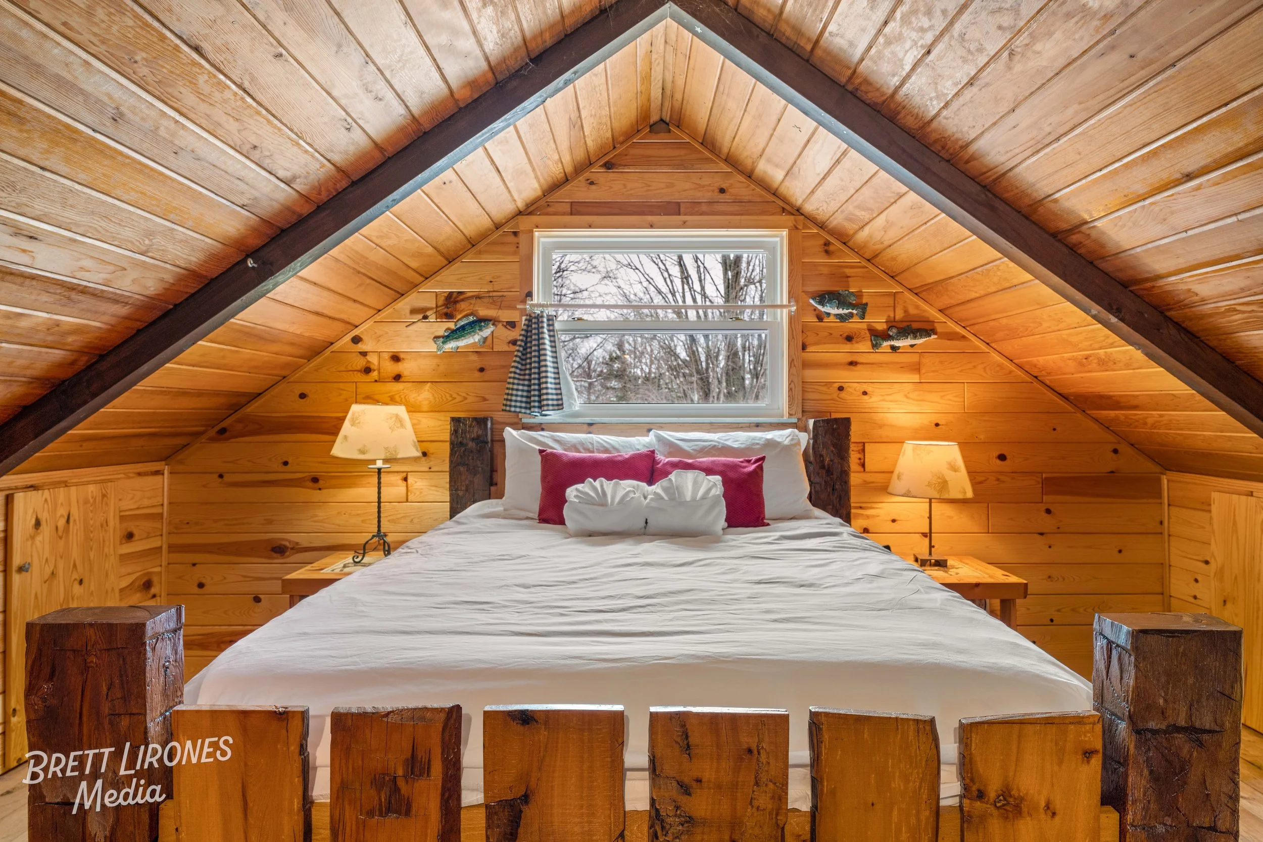 A rustic bedroom with a wooden sloped ceiling and walls, a bed with white linens and red pillows, nightstands with lamps, and a window with curtains. Fish decorations are on the wall.