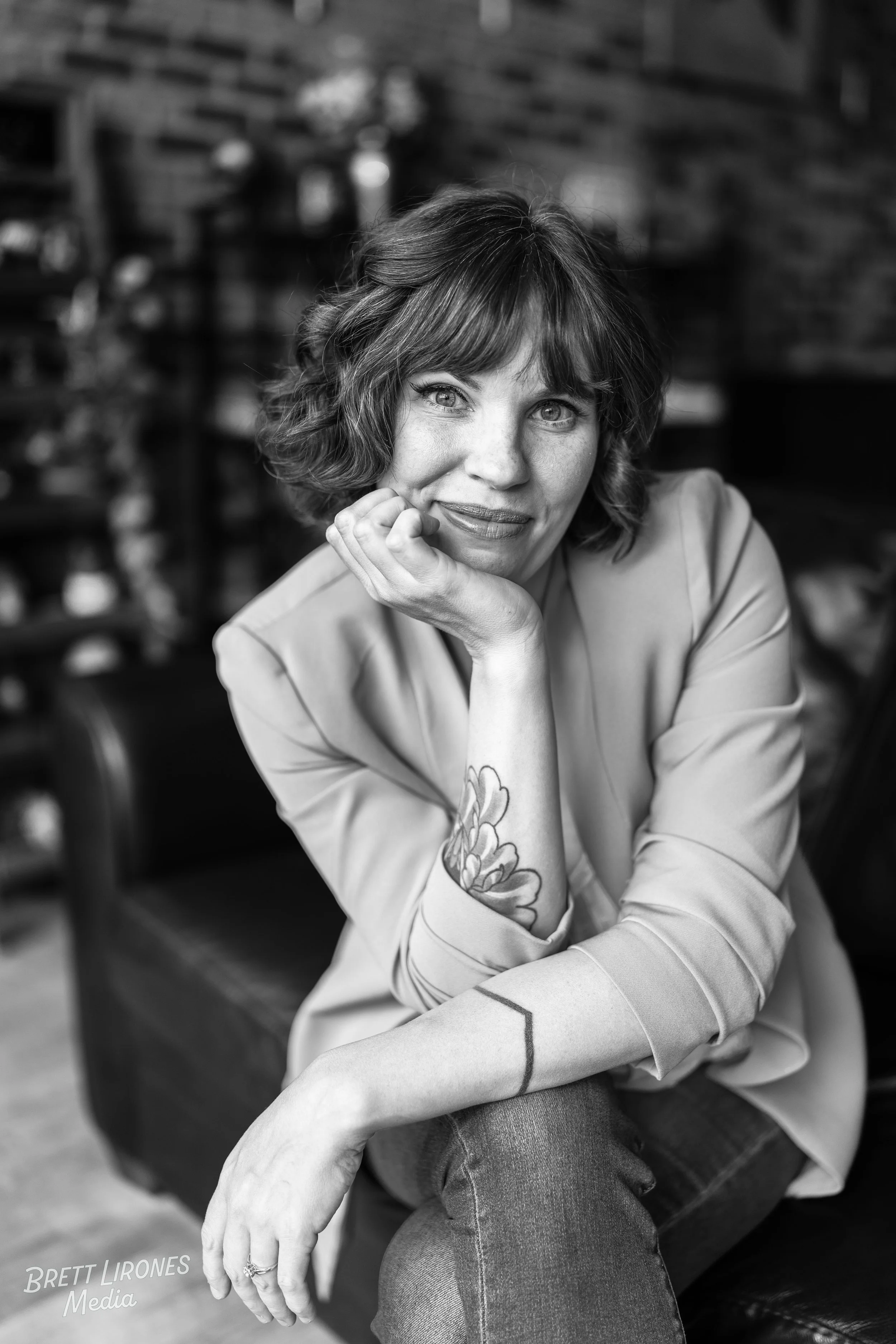 Black-and-white portrait of a woman with short wavy hair, resting her chin on her hand, sitting indoors with shelves in the background.