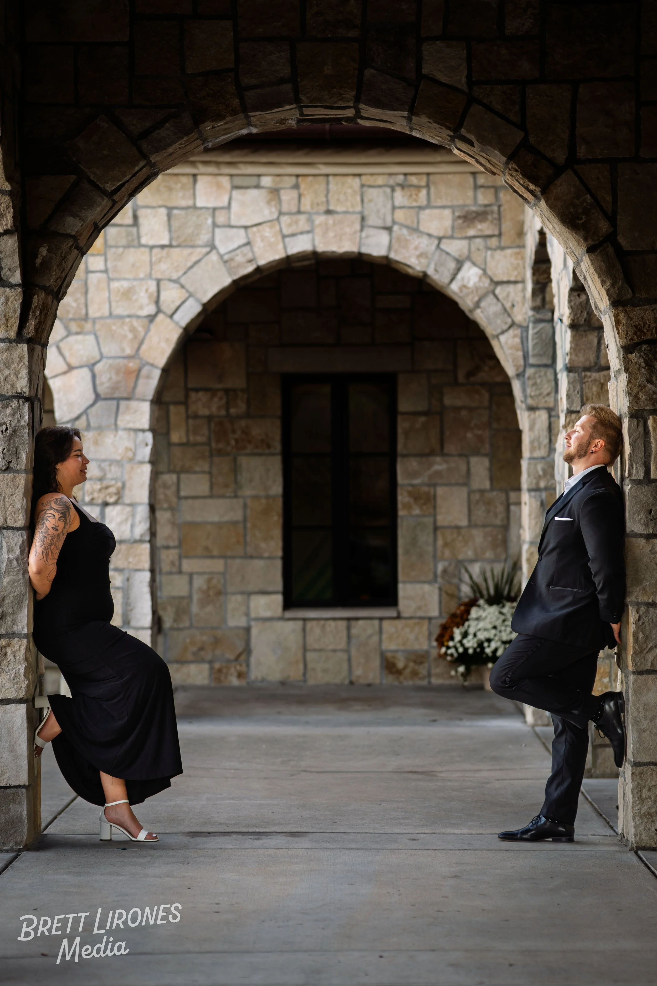 A woman and a man are leaning against stone walls in an archway, facing each other and smiling during a romantic moment.