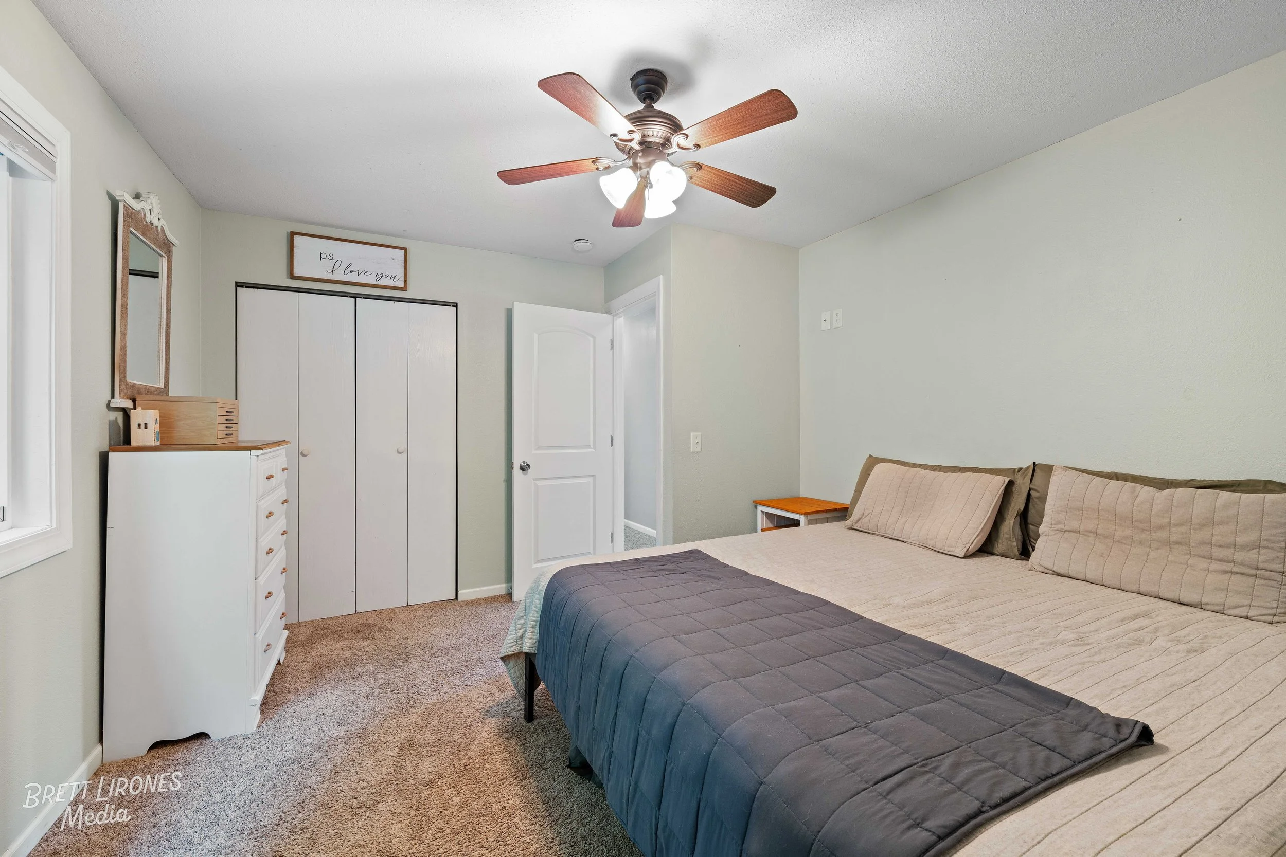 A simple bedroom with a bed, nightstand, dresser, mirror, closet, ceiling fan, and a wall sign that reads 'P.S. I love you.'