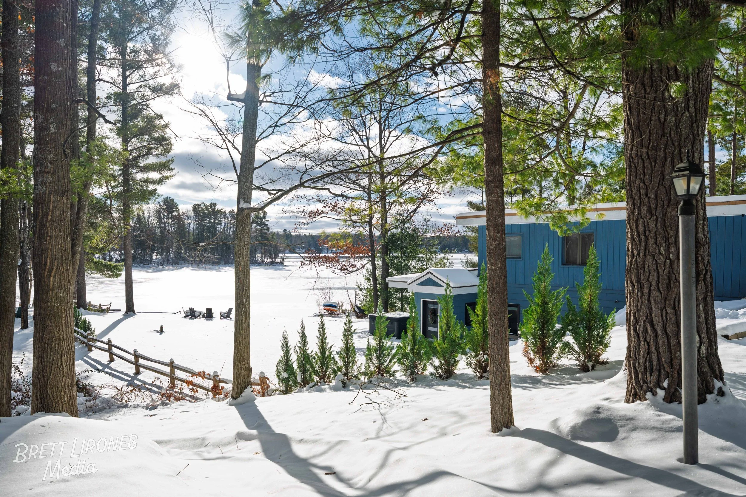 Snow-covered backyard with trees, a blue house, and Adirondack chairs near a frozen lake on a partly cloudy day.