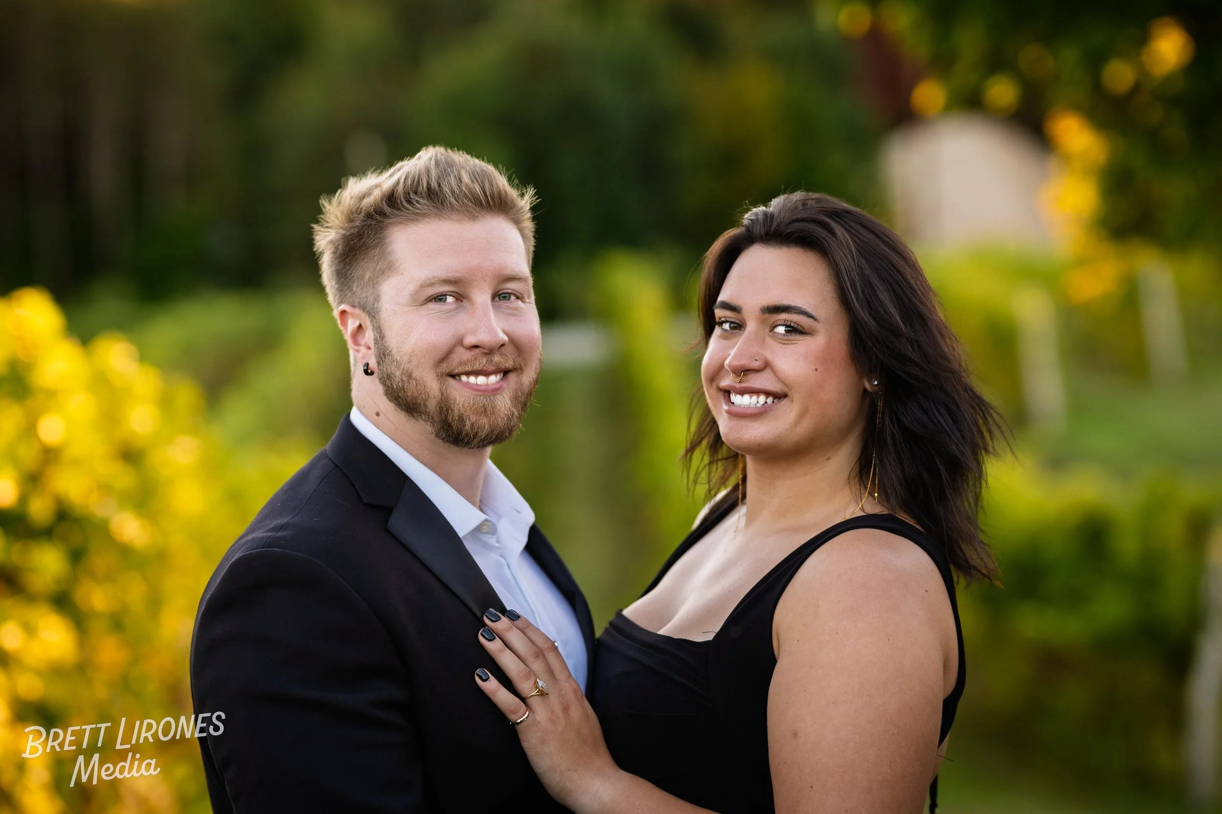 A couple smiling outdoors with greenery and trees in the background, the man has blonde hair and a beard, wearing a black suit jacket and white shirt, and the woman has dark hair, wearing a black dress and jewelry.