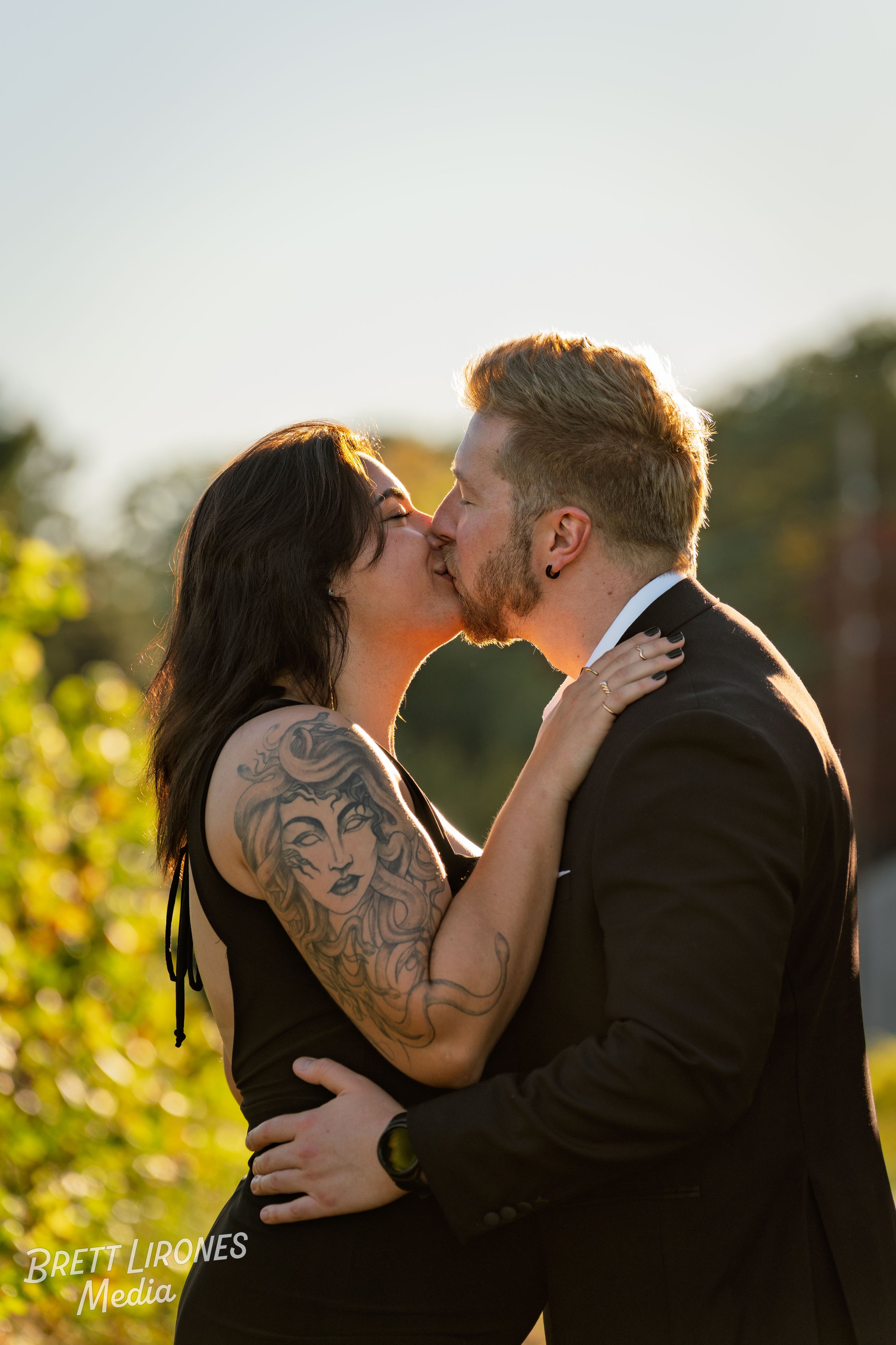 A couple kissing outdoors in the sunlight, with the woman having tattoos on her arm and wearing black, and the man in a black suit, embracing each other.