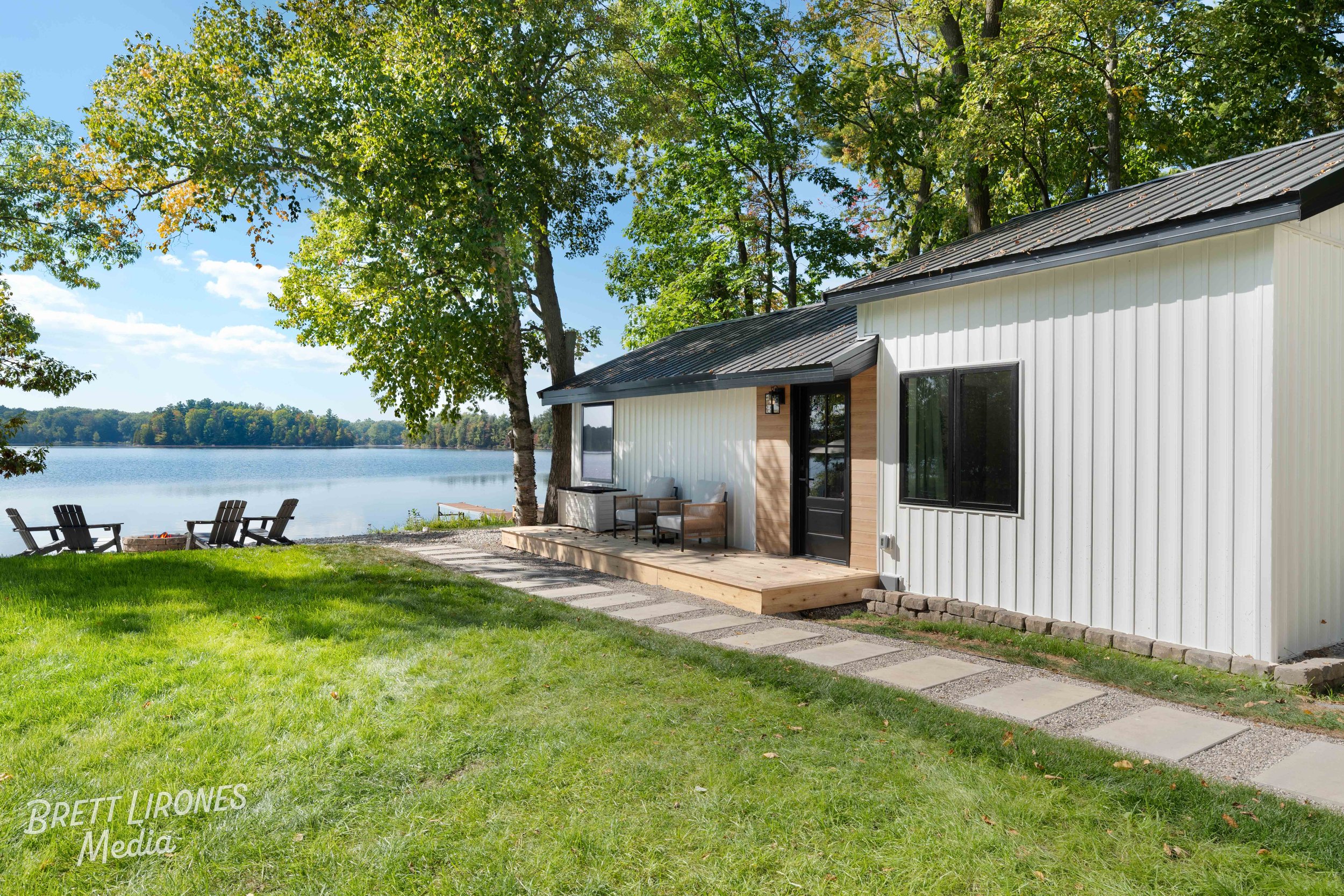 Modern white house with black trim, outdoor seating area on a wooden deck, lake view with trees, Adirondack chairs on grass, clear sky.