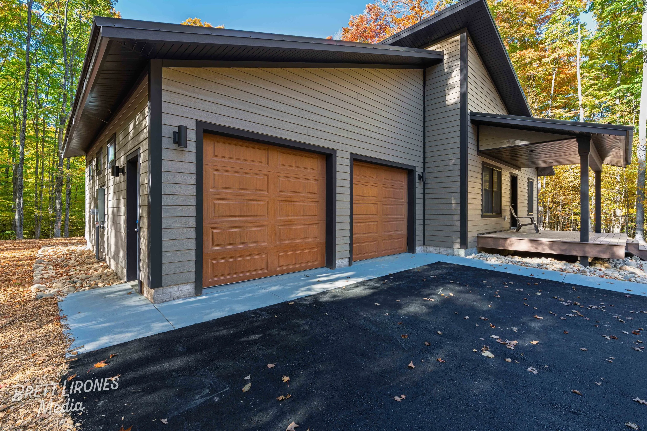 Exterior view of a modern house with a two-car garage featuring wooden doors, surrounded by trees with fall foliage, and a small porch with a chair.