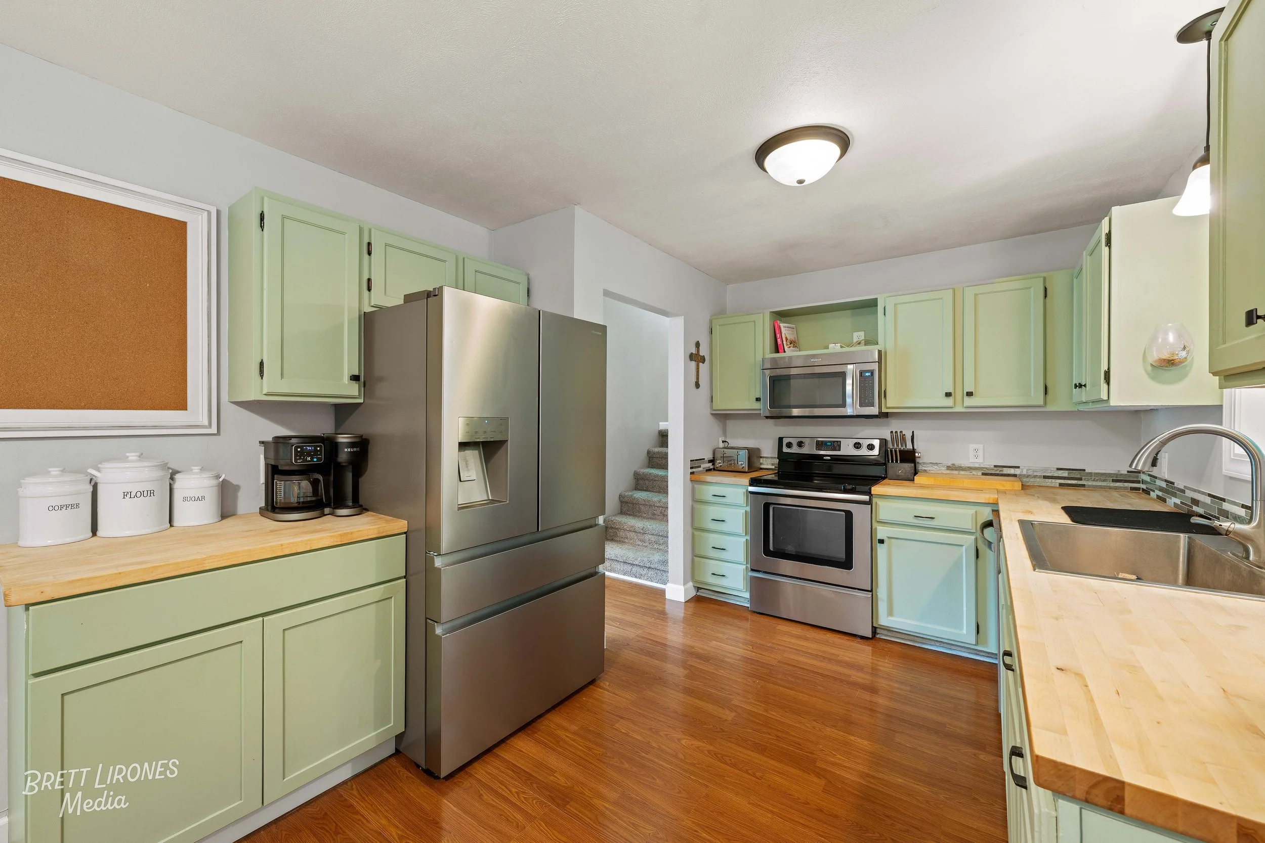 A kitchen with light green cabinets, stainless steel refrigerator, black appliances, wooden countertops, and hardwood floor.