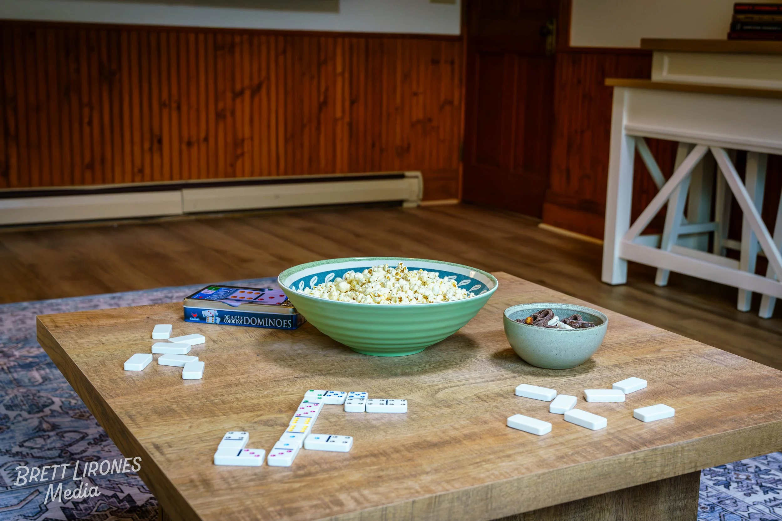 A wooden coffee table with scattered domino tiles, a bowl of popcorn, and a smaller bowl with chocolates, in a living room with wood-paneled walls and bookshelves.