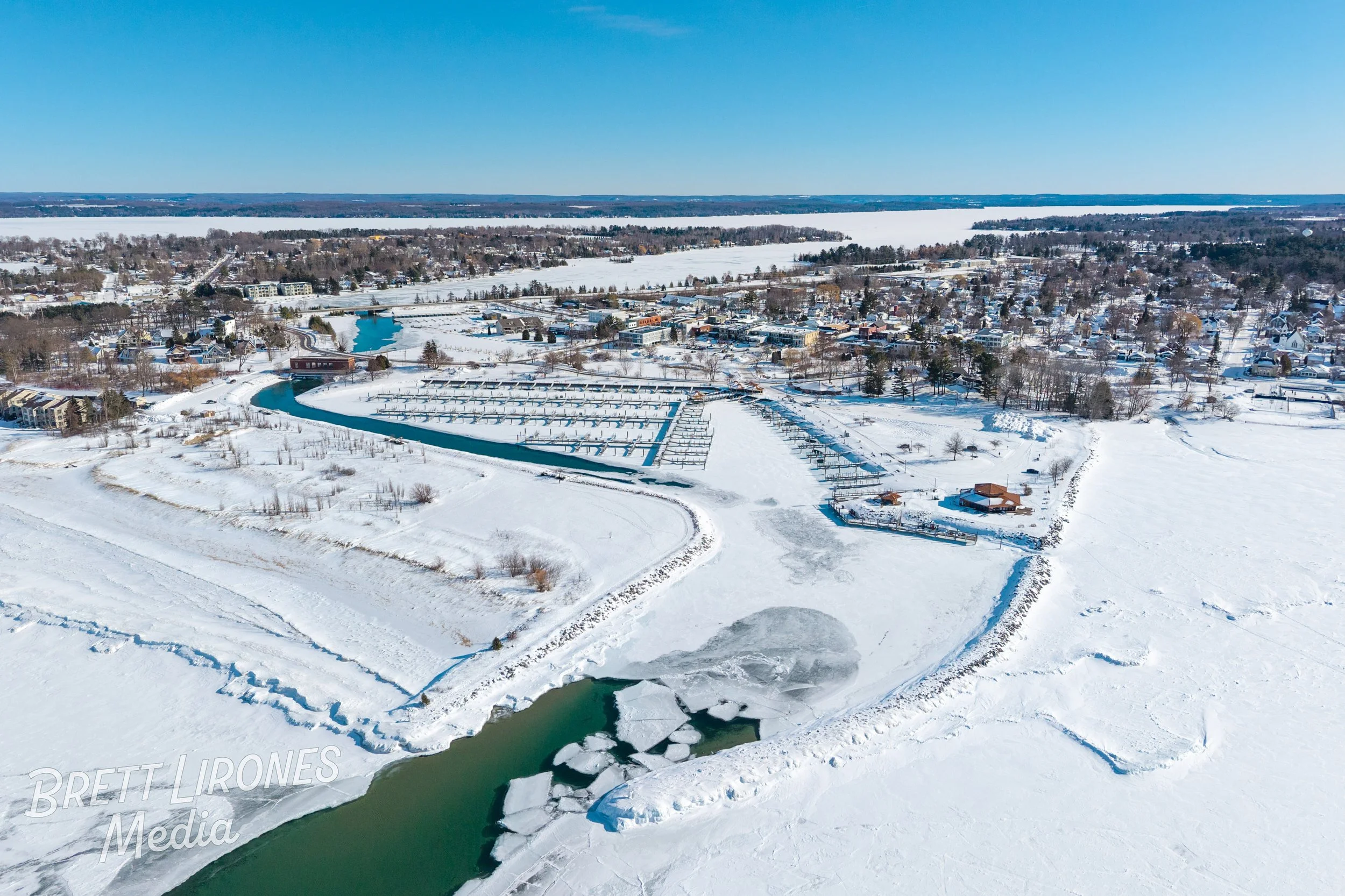 Aerial view of a snow-covered landscape featuring a frozen body of water with ice formations, and a marina with boats docked along a winding canal. Residential houses and trees are visible in the background under clear blue skies.