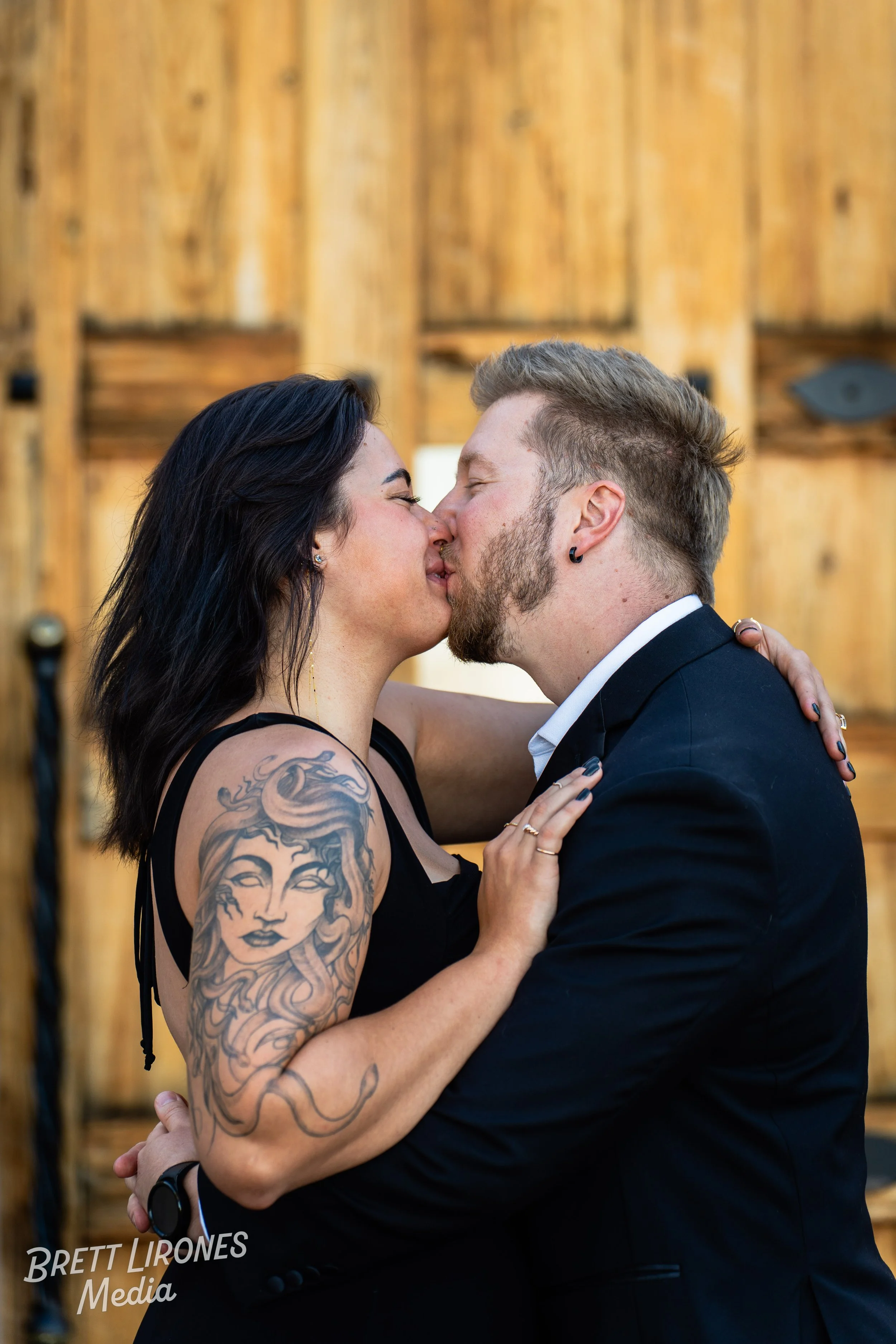 A couple kissing, with the woman having dark hair, tattoos on her arm, and wearing a black dress. The man has light hair, a beard, earrings, and is wearing a black suit. They are close together with the woman's hand on the man's shoulder, against a w