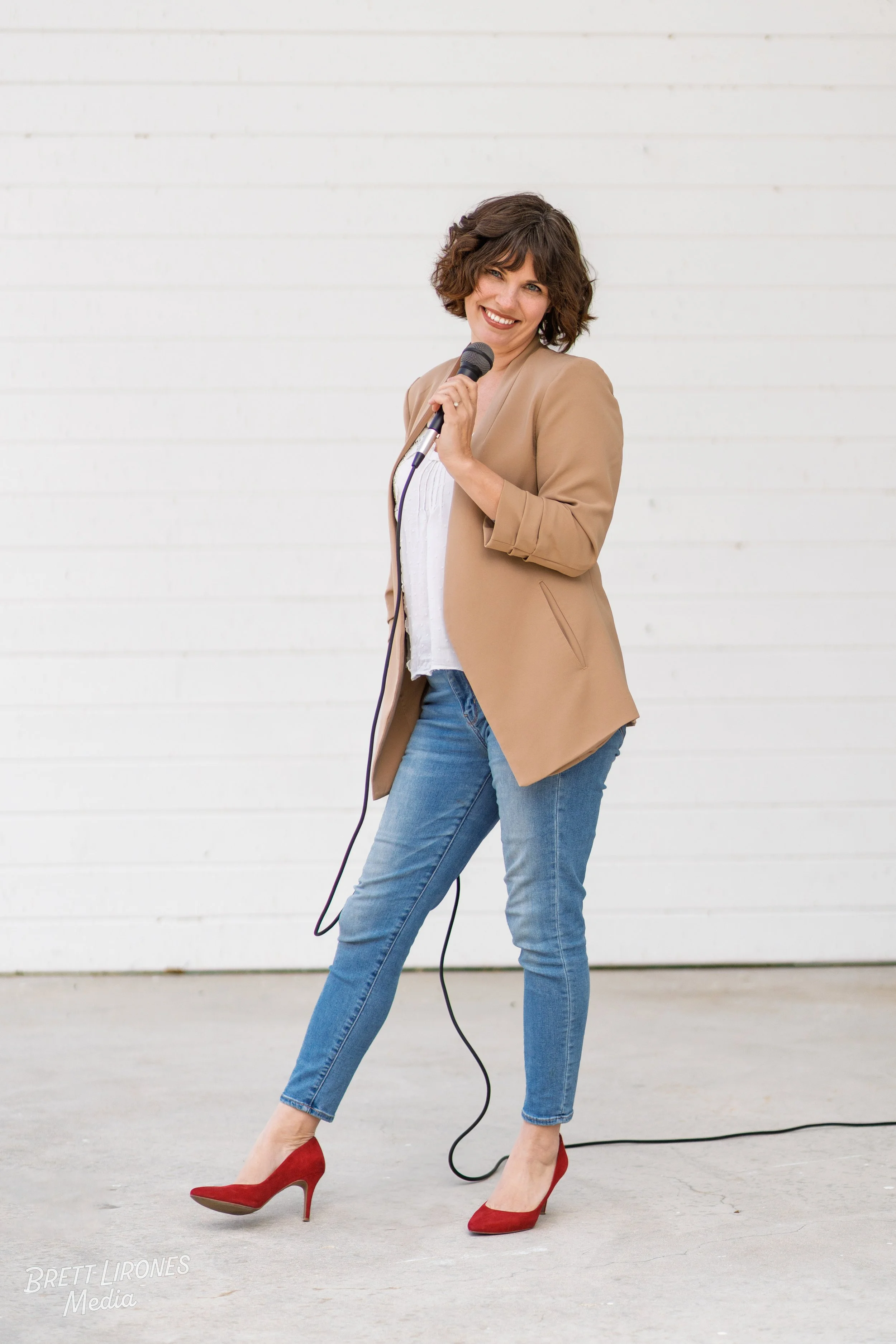 A woman with short curly brown hair is standing in front of a white wall, holding a microphone, and smiling at the camera. She is wearing a tan blazer, a white top, blue jeans, and red high heels.