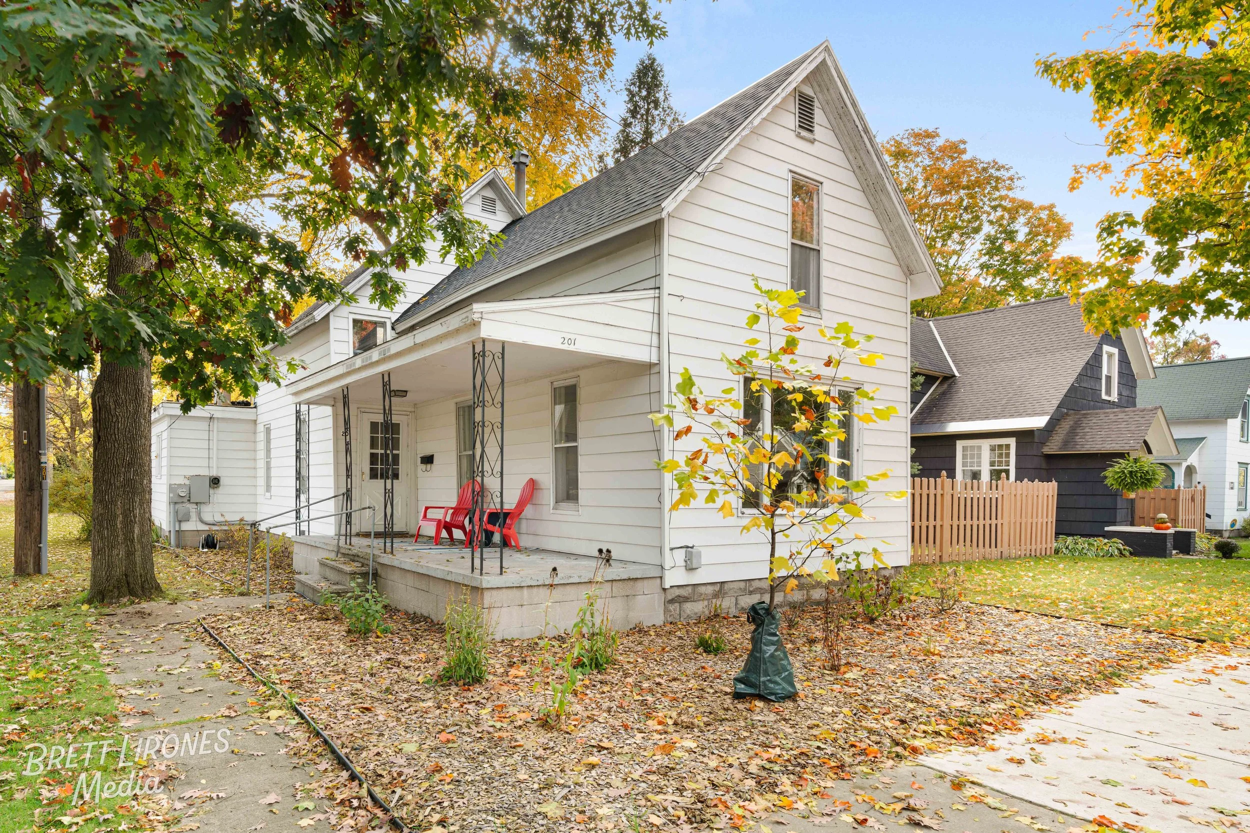 Front view of a white house with a small covered porch, two red chairs on the porch, and a young tree with yellow leaves in front. Surrounding trees have autumn foliage, and there are neighboring houses with different colored exteriors. Fallen leaves