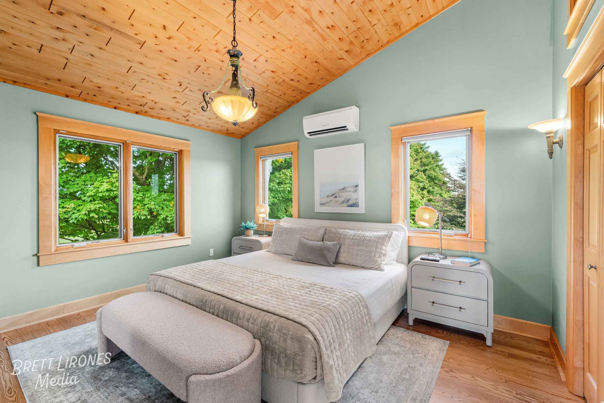Cozy bedroom with green walls, wooden ceiling, and three large windows revealing green trees outside. The room has a white bed with beige bedding, two bedside tables with lamps and decor, and a curved bench at the foot of the bed. There's a chandelie