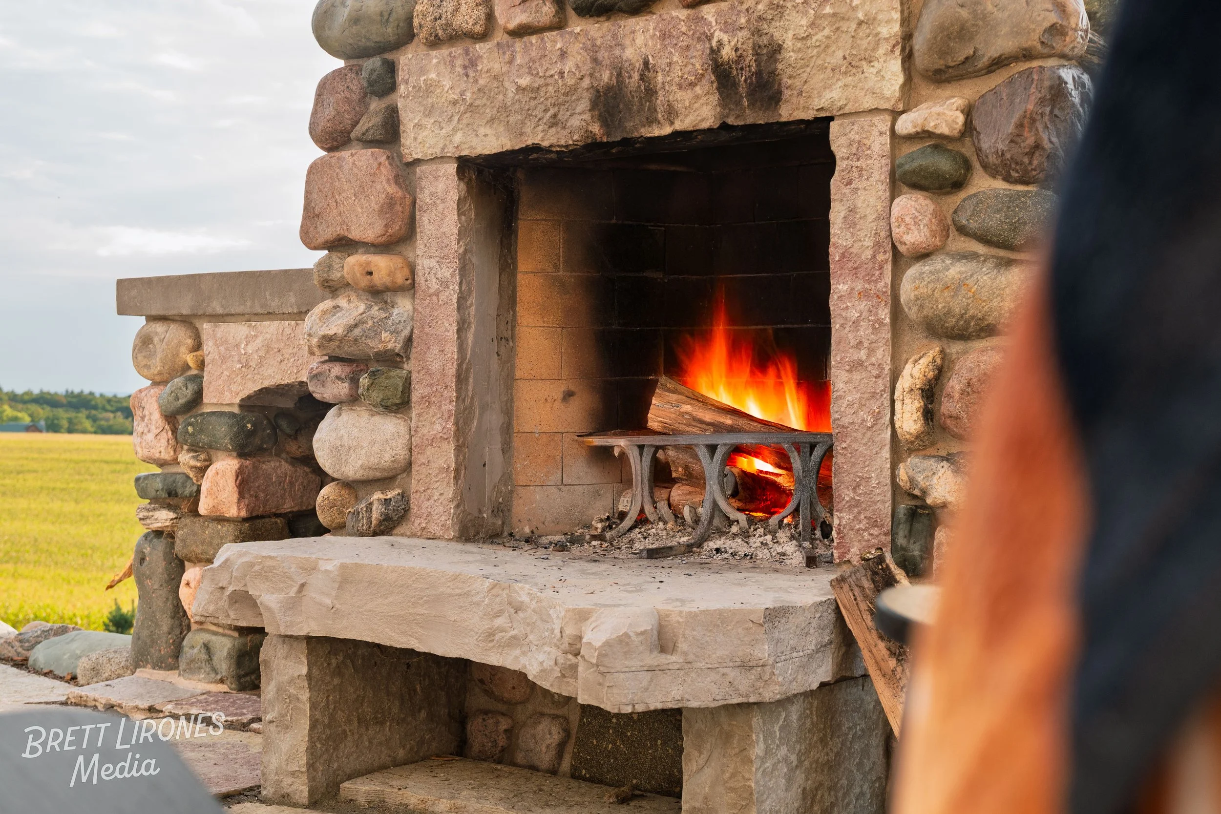 An outdoor stone fireplace with a burning fire and logs inside, with a grassy field and cloudy sky in the background.