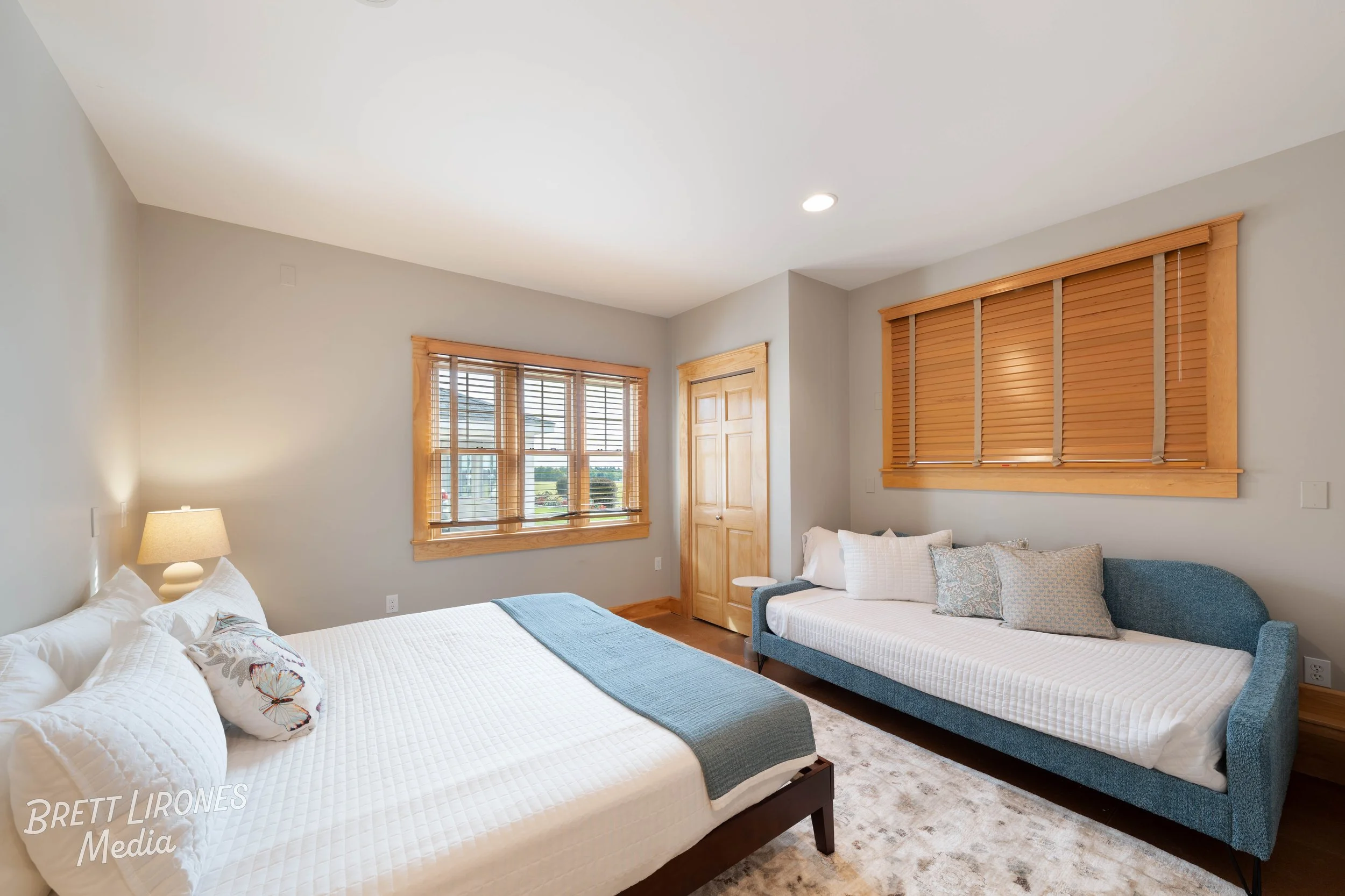 Bedroom with two beds, one large bed with white bedding and a blue blanket, and a blue daybed with several pillows, two windows with wooden blinds, light-colored walls, hardwood floors, and a rug.
