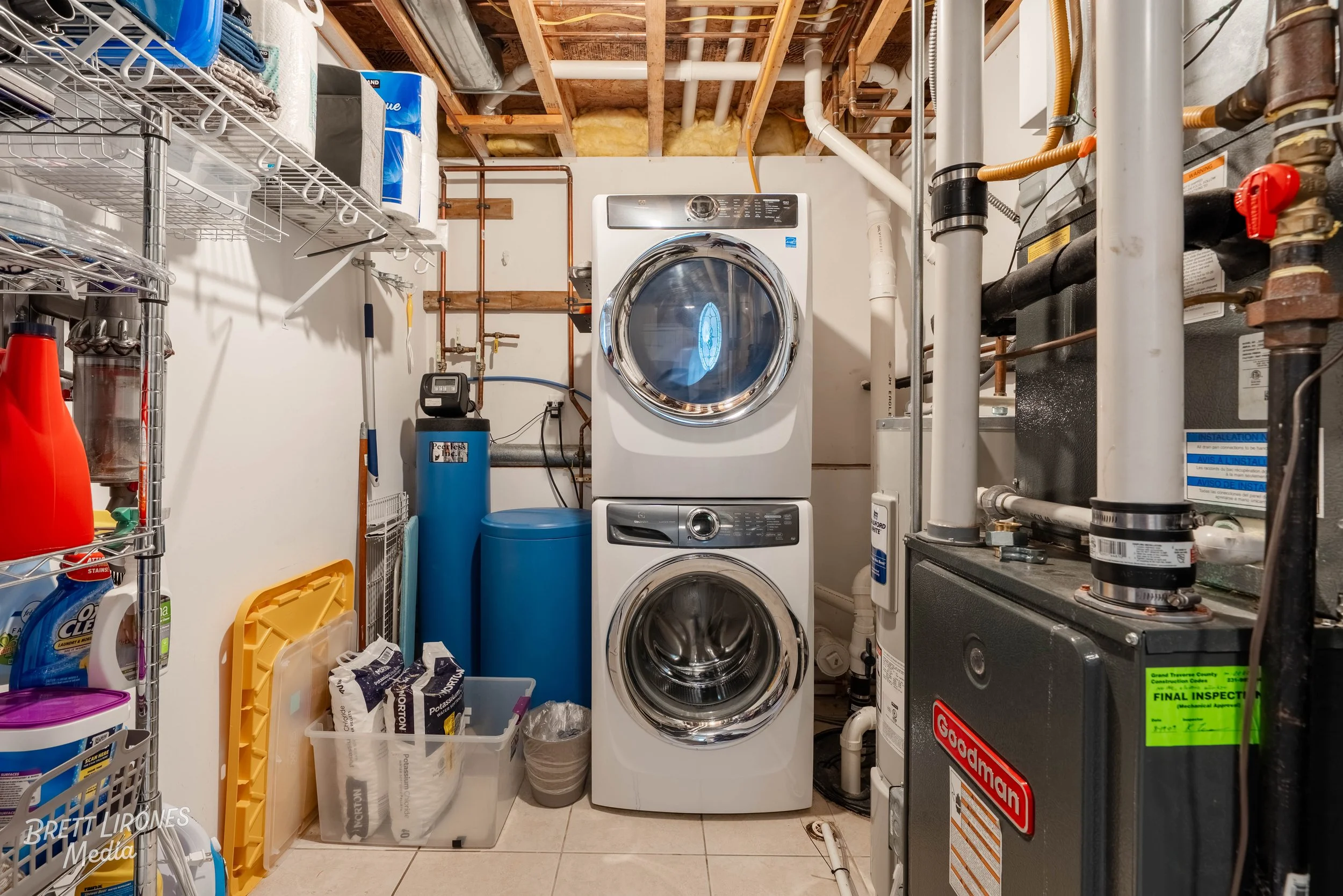 Laundry room with a white stacked washer and dryer, various laundry supplies on a metal shelf, a blue water filter and softener, and a black Goodman furnace to the right, with exposed ceiling pipes and insulation.