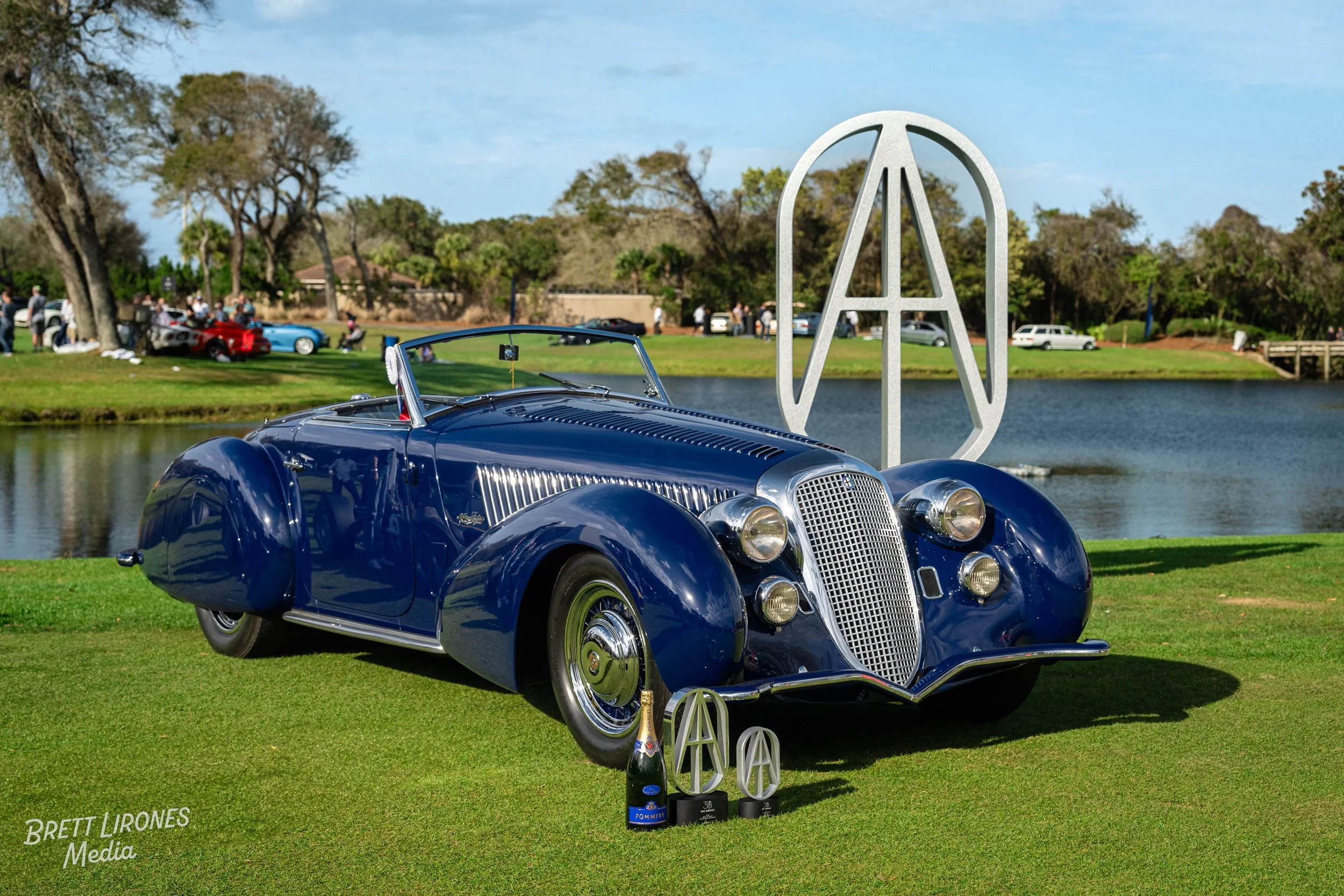 A vintage blue car with chrome accents parked on grass near a body of water, with a large peace sign sculpture behind it and several trophies and a bottle of champagne in front, at an outdoor event with people and other classic cars in the background