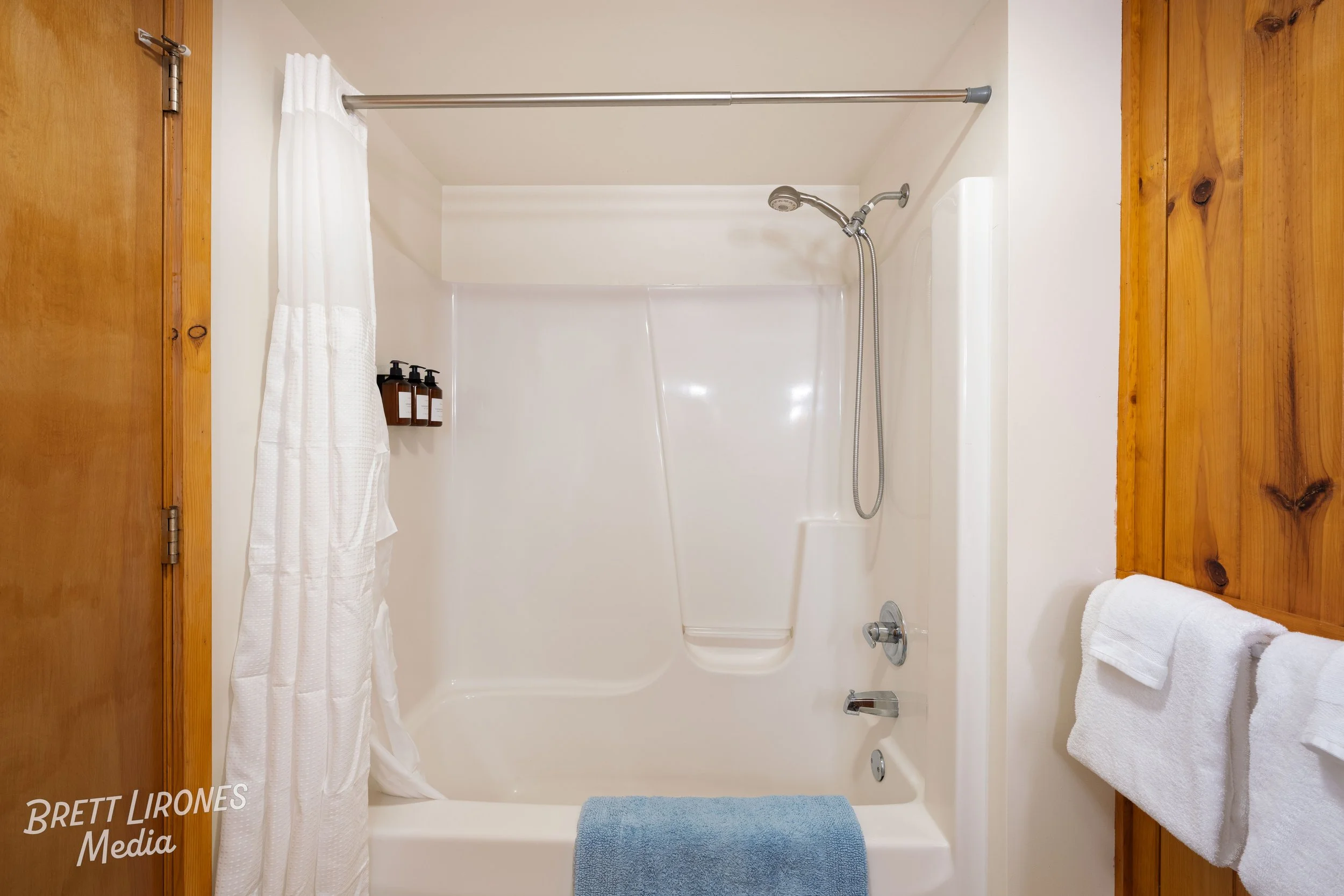 Bathroom shower area with white walls, a curved shower rod with a white curtain, bottles of soap or shampoo on a wall shelf, and brown wooden paneling on the right side with white towels hanging on a bar.