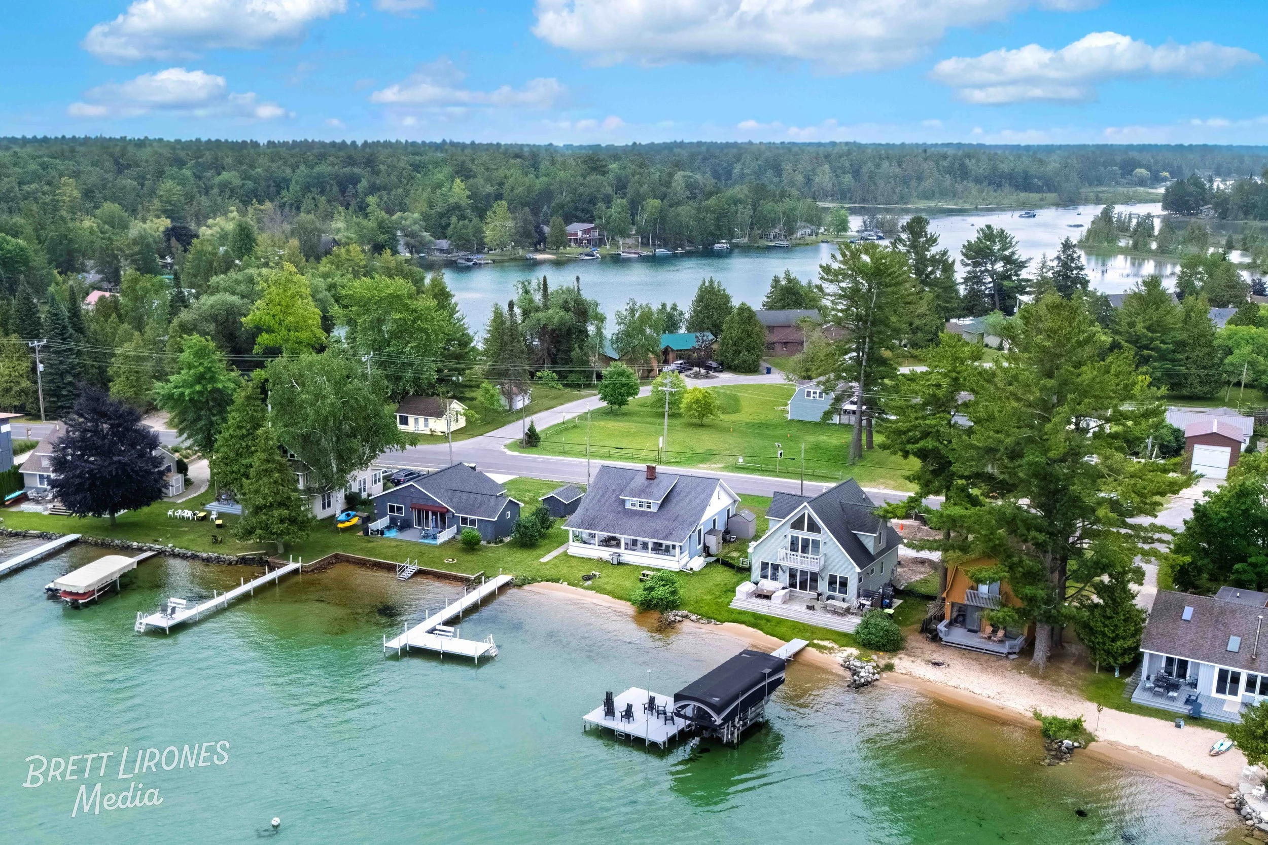 Aerial view of a lakeside residential area with houses, docks, and boats on a green lake, surrounded by a dense forest under a partly cloudy sky.