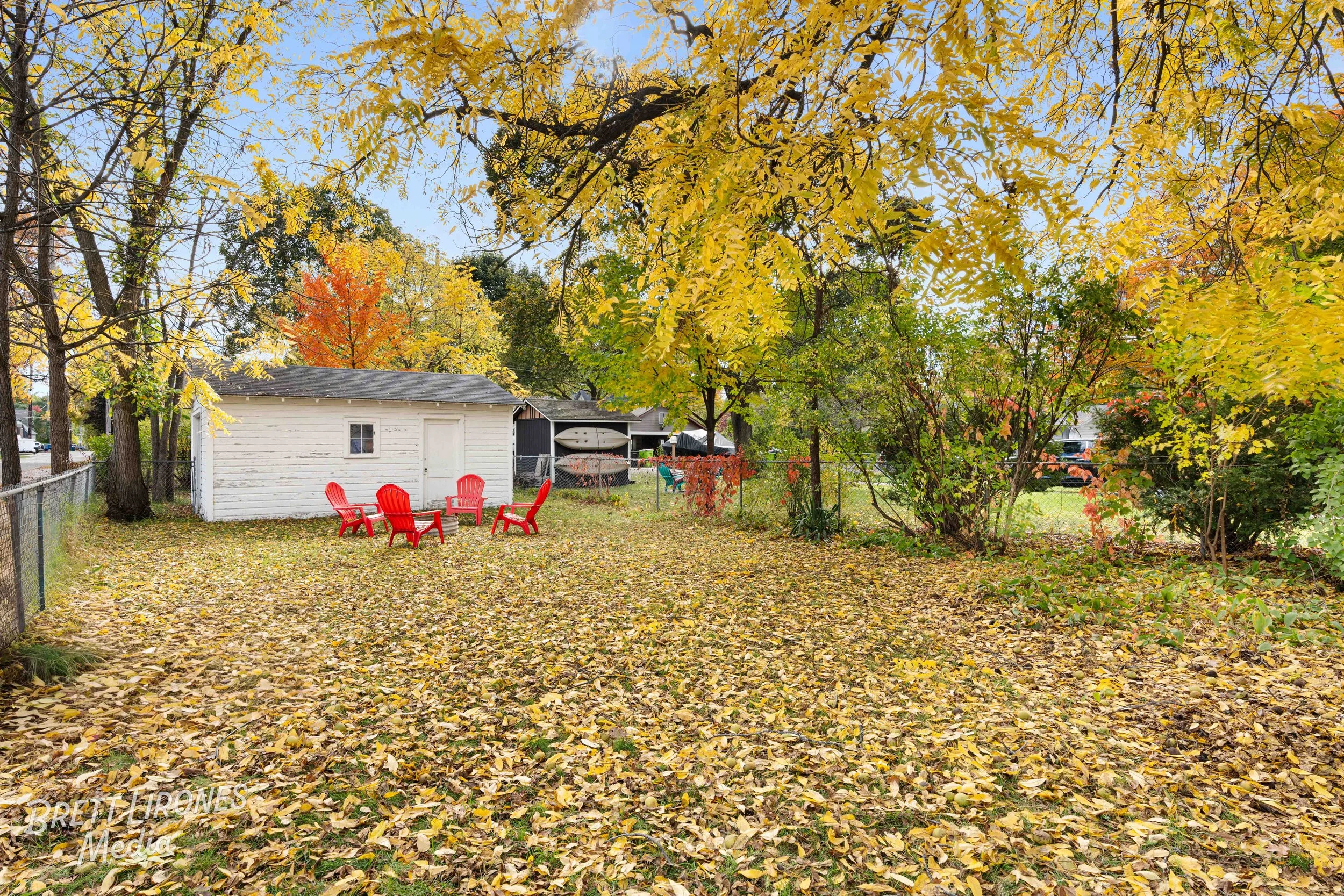 Backyard with fallen leaves, a white shed, and four red chairs around a small table, surrounded by trees with autumn leaves.
