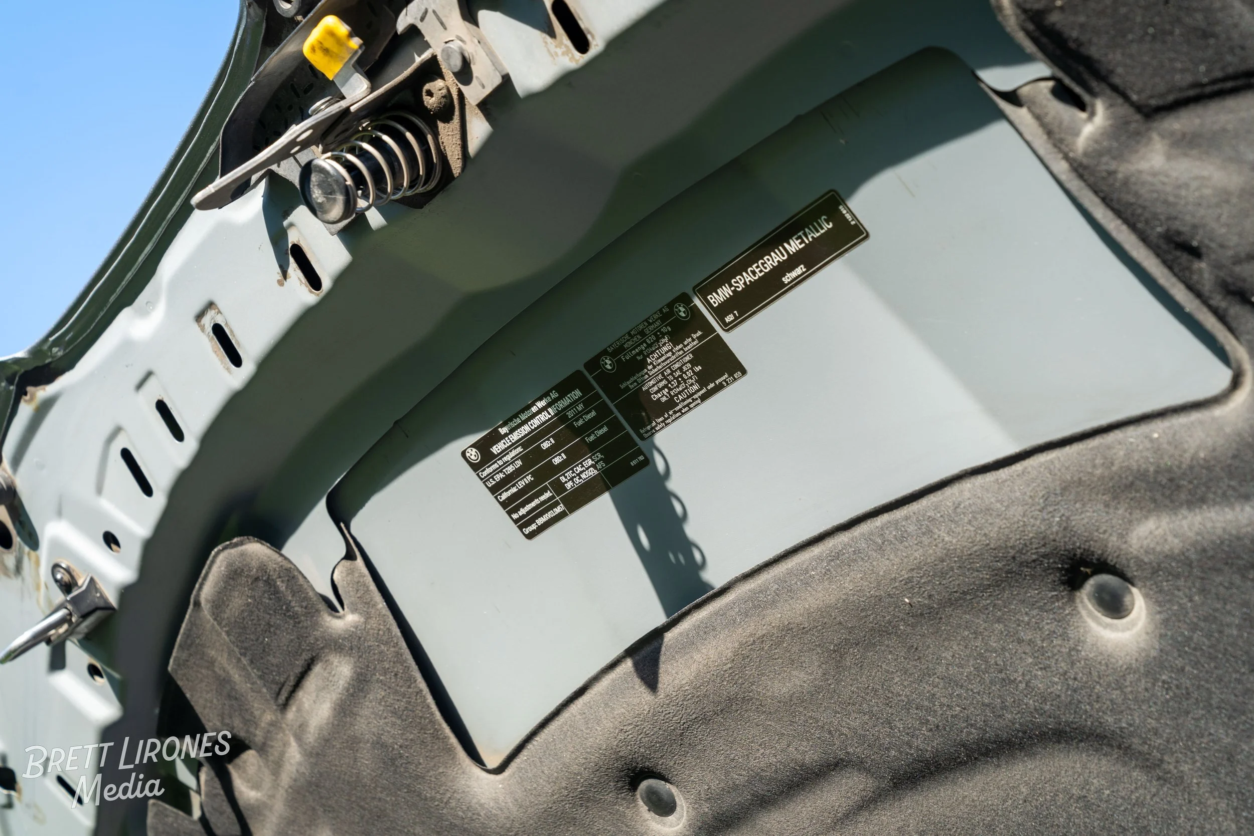 Close-up of a vehicle's interior showing a metal box with black labels, including one that reads 'BMW-SPACEGRAY METALLIC,' surrounded by black fabric and a spring mechanism on the top left corner.