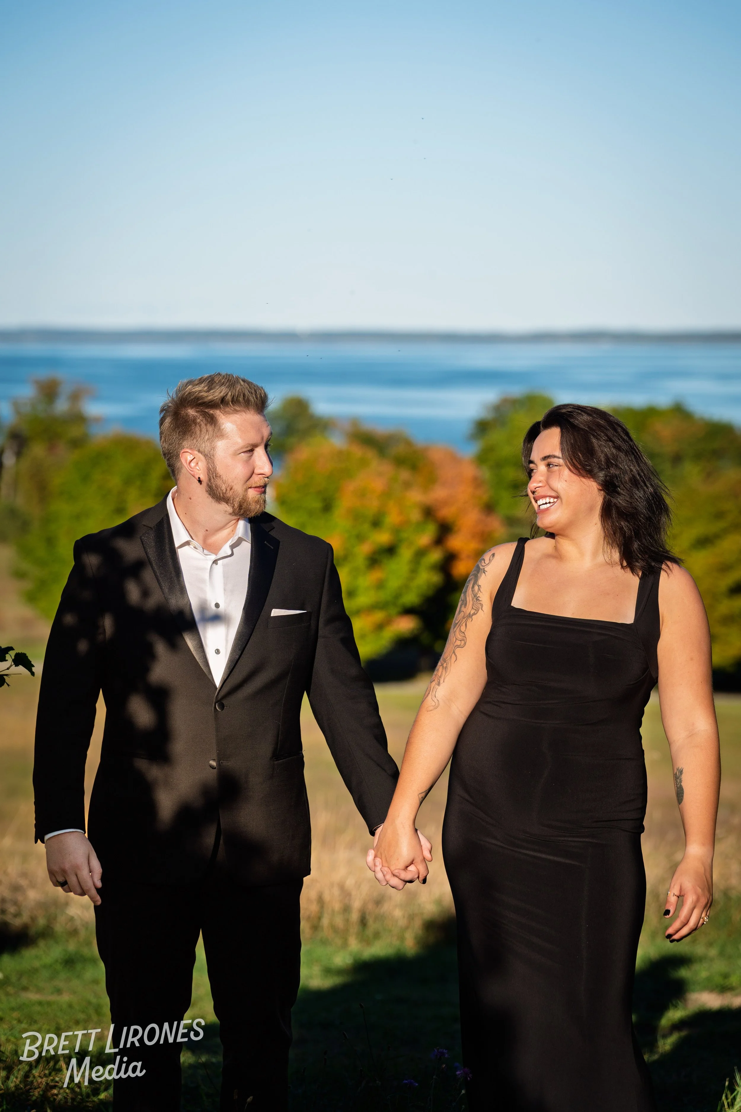 A couple holding hands, walking outdoors near a lake, with trees and a clear blue sky in the background.