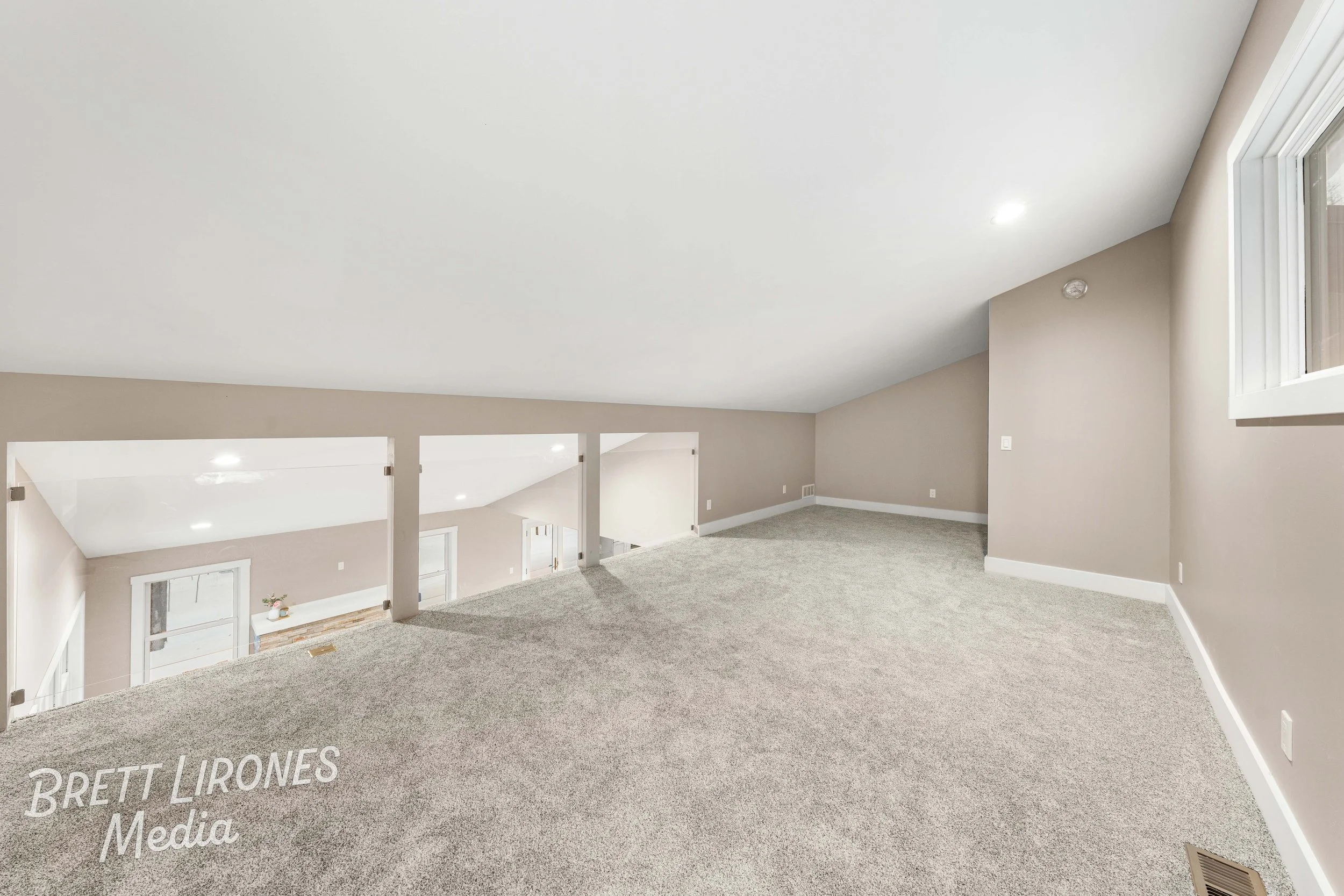 Empty finished basement with beige walls, gray carpet, and a white ceiling, overlooking the lower level via a half wall with open spaces, and a window on the right side.