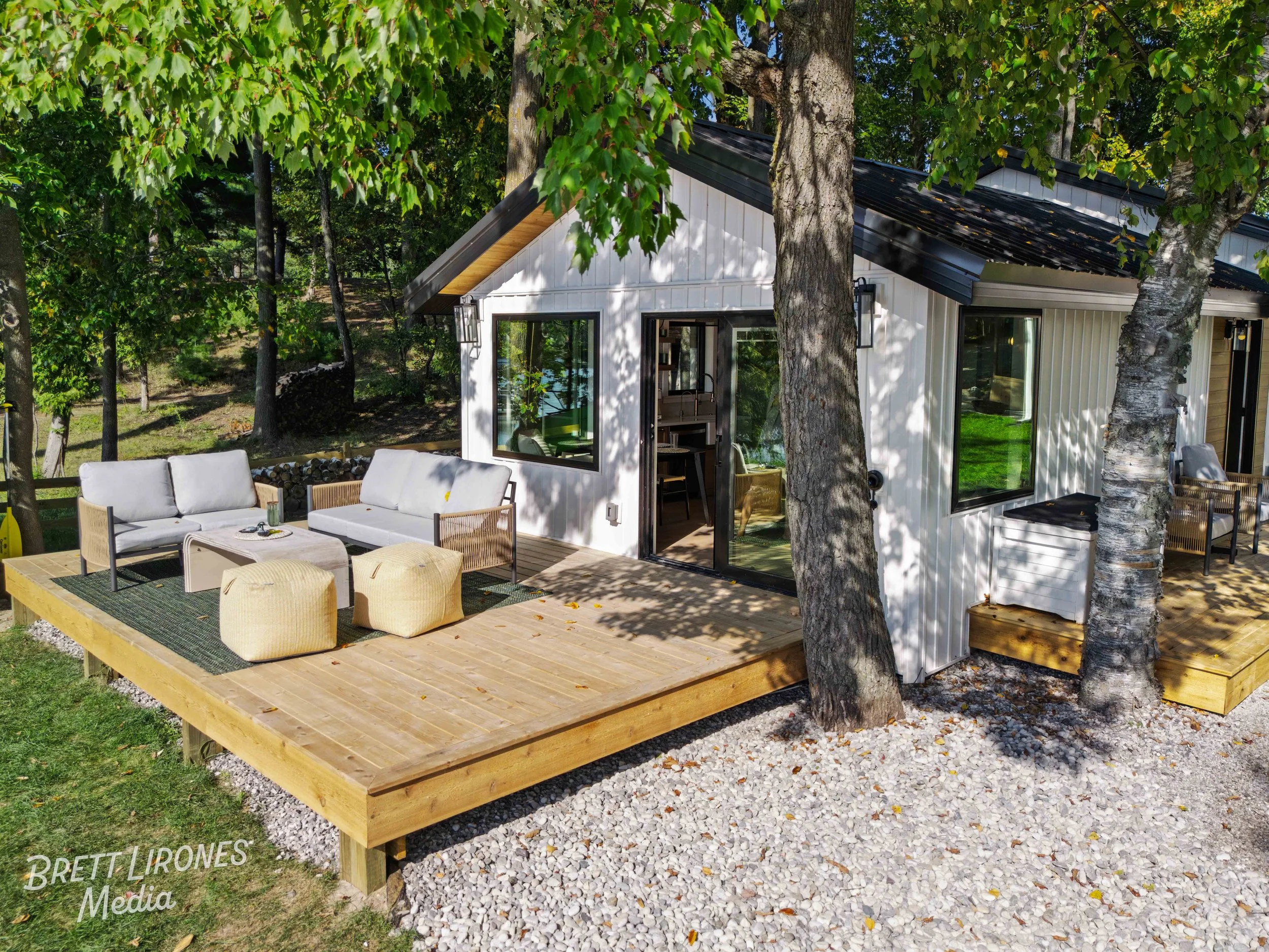 Exterior view of a modern white house with black roof, large windows, and a wooden deck. The deck has outdoor seating with sofas and ottomans, surrounded by trees and greenery.