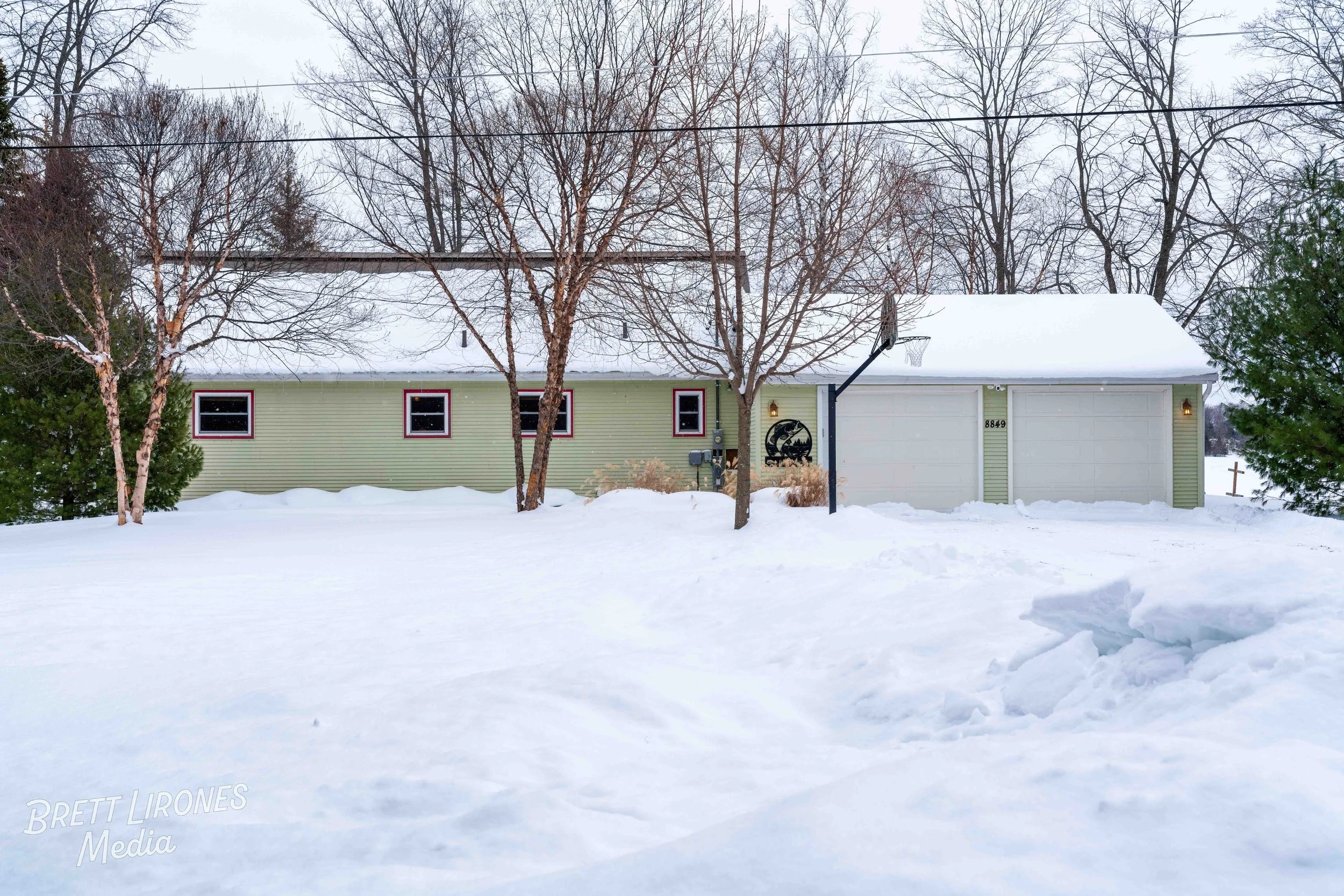 Snow-covered house with green siding, three small windows, and an attached garage. Leafless trees in front and snow on the ground, winter setting.
