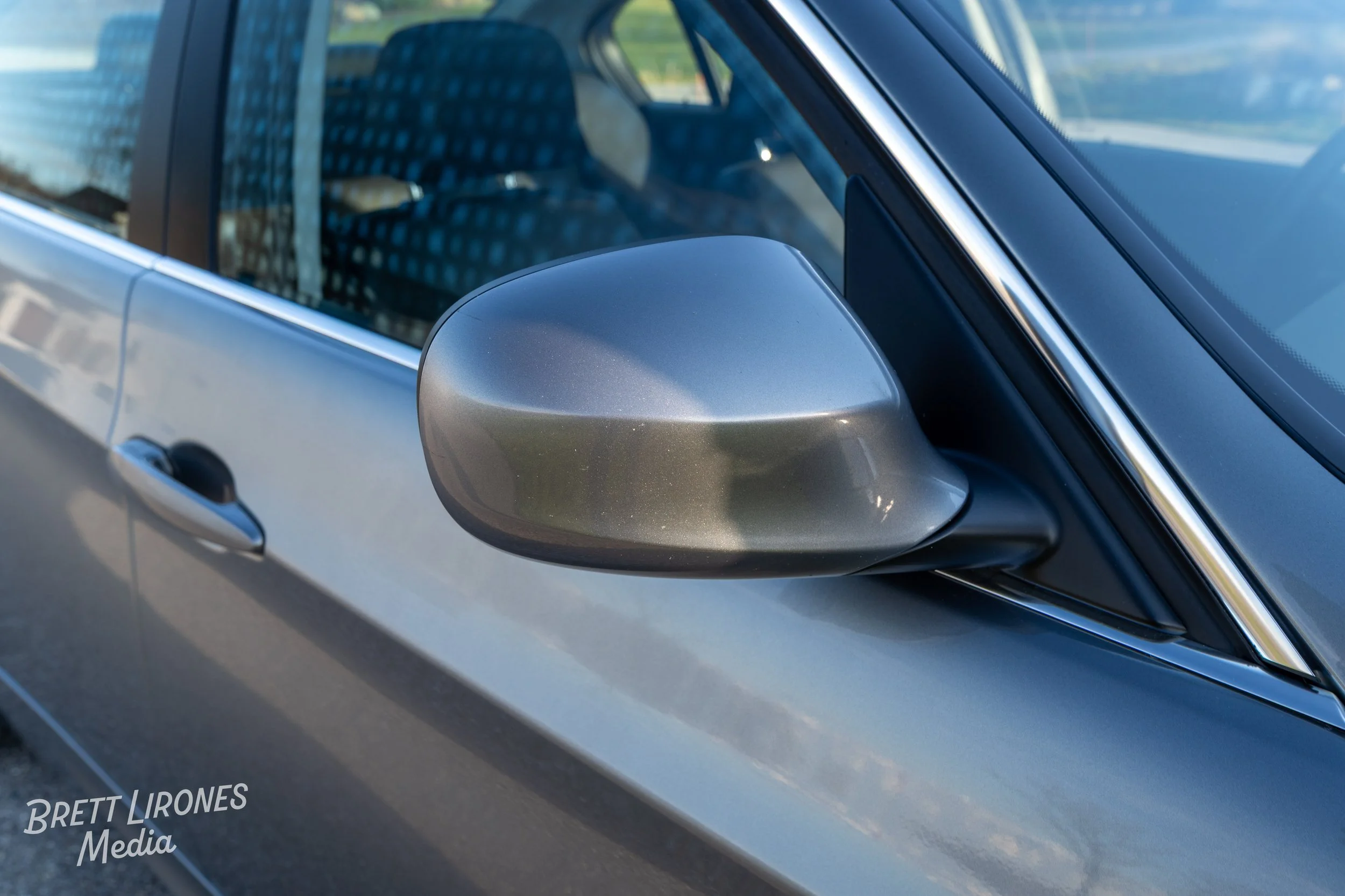 Close-up of a silver car's side mirror and part of the front door, with a black window trim and a window showing a patterned interior seat.