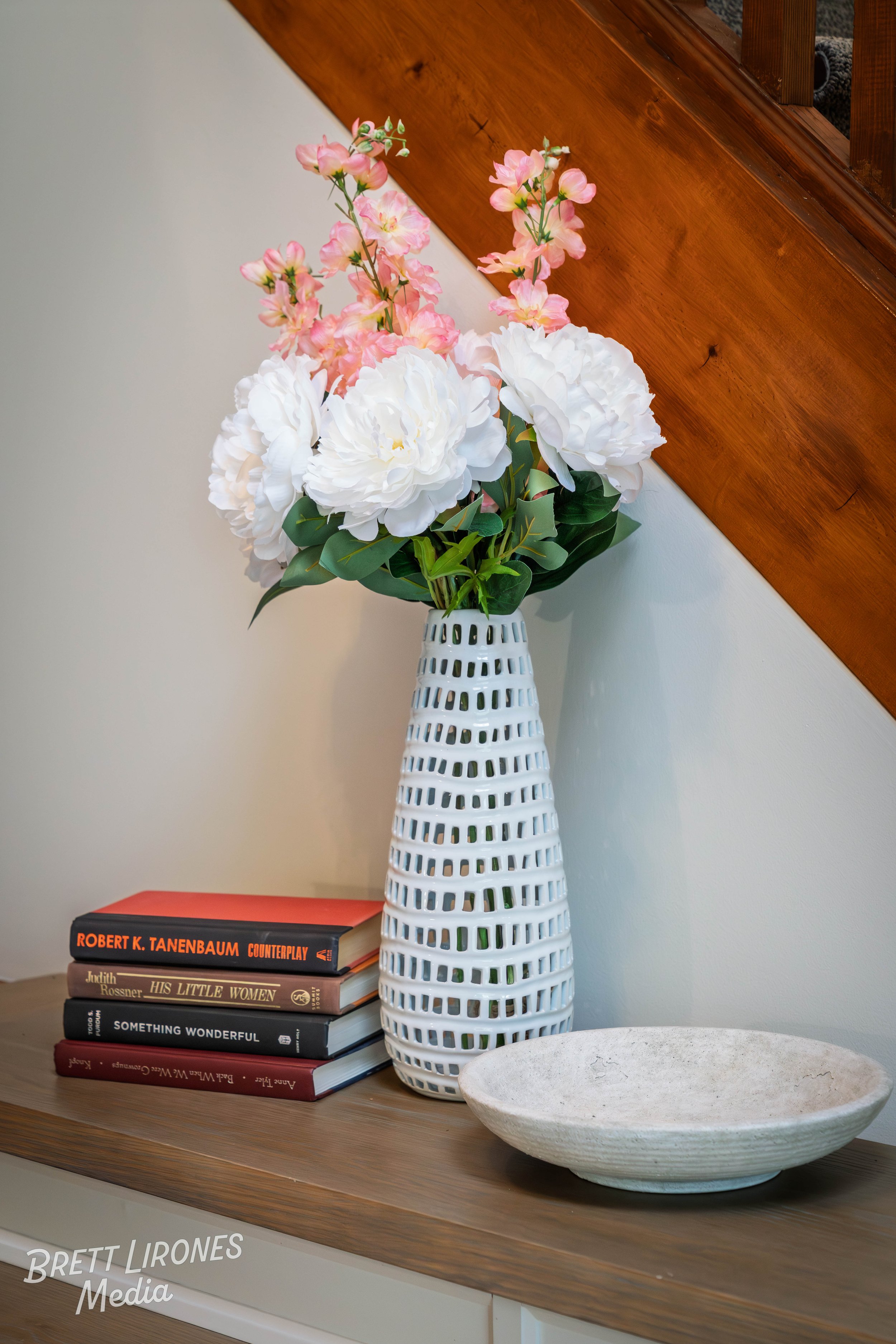 A white vase with a lattice pattern holding pink and white flowers sits on a wooden shelf next to a stack of books and a large white bowl. There is a wooden staircase with a carpeted runner in the background.