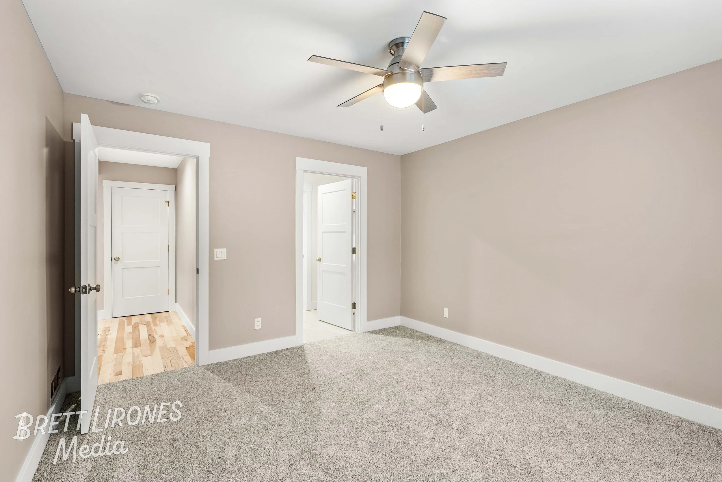 Empty room with beige walls, gray carpet, white trim, a ceiling fan with lights, and two open doorways leading to other rooms.