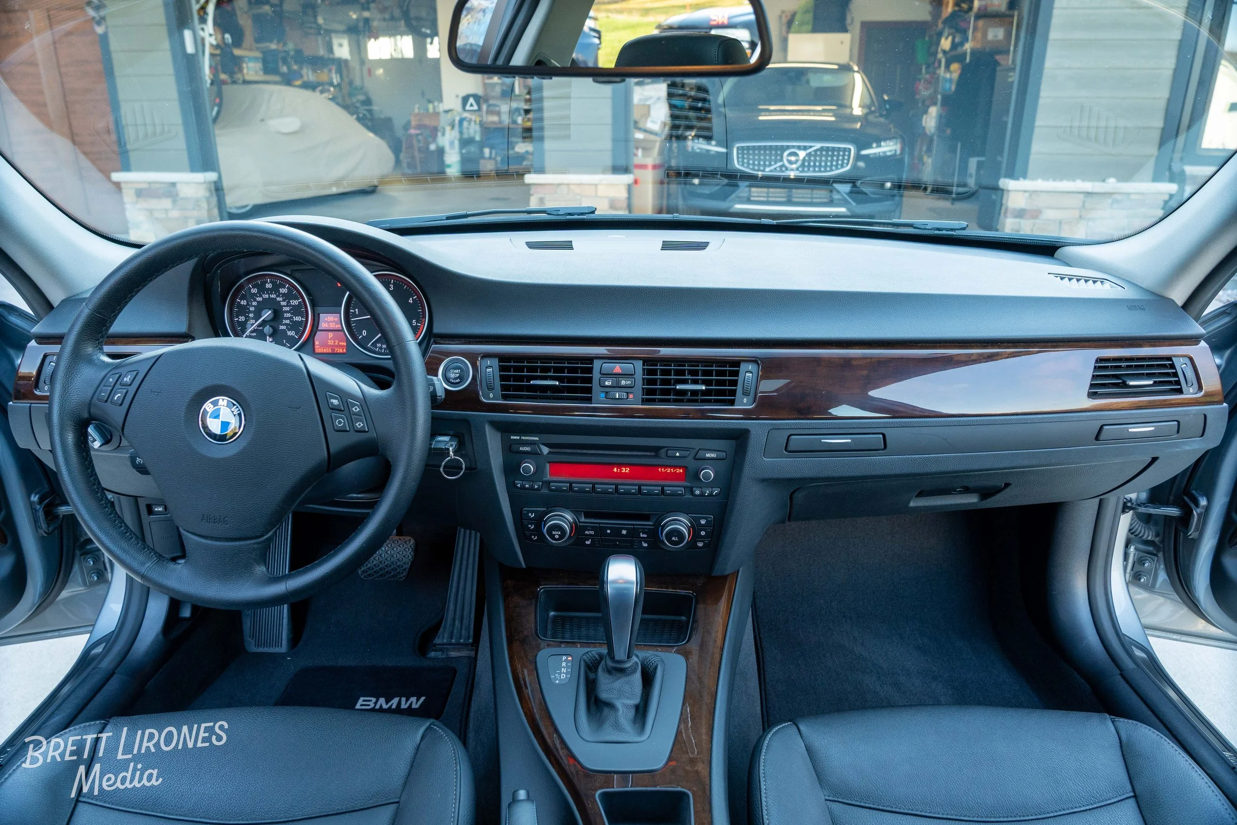 Interior of a BMW car, showing the steering wheel, dashboard, gear shifter, and front seats, with a view through the windshield of another vehicle and garage area outside.