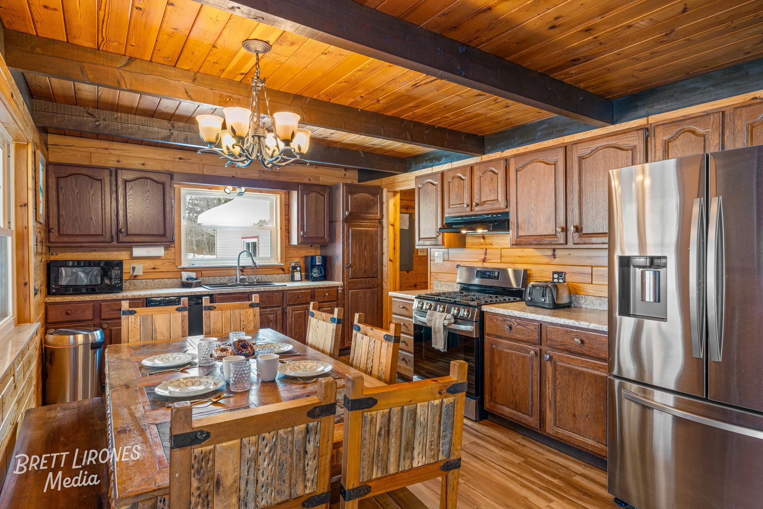 Rustic kitchen with wooden cabinets, a wood-paneled ceiling, stainless steel appliances, a dining table set with plates and cups, and a chandelier