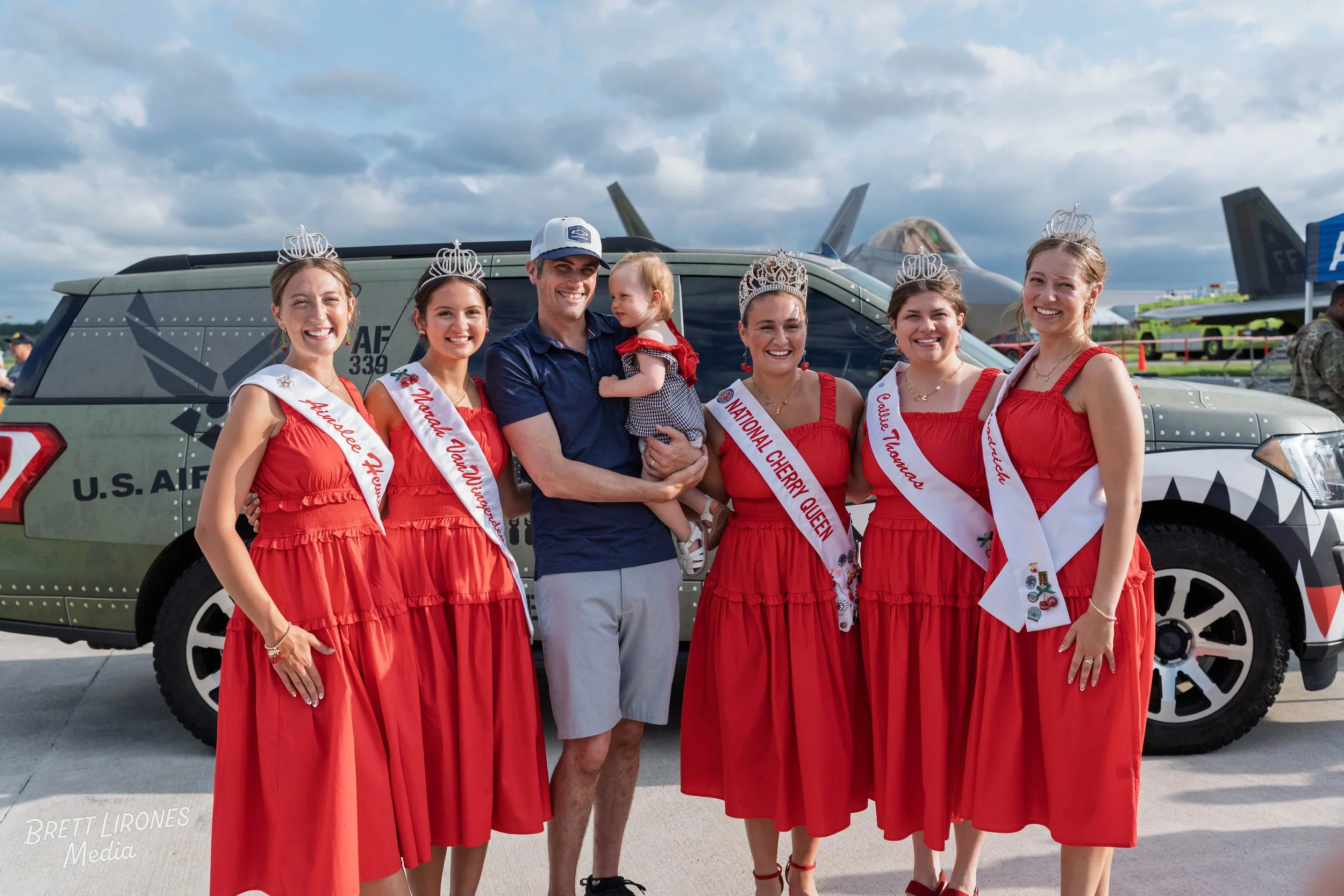 Five women wearing red dresses and sashes with titles such as 'Alabama Queen' and 'National Cherry Queen,' standing next to a man holding a young girl, in front of a camouflage-patterned U.S. Air Force vehicle, with military jets in the background.