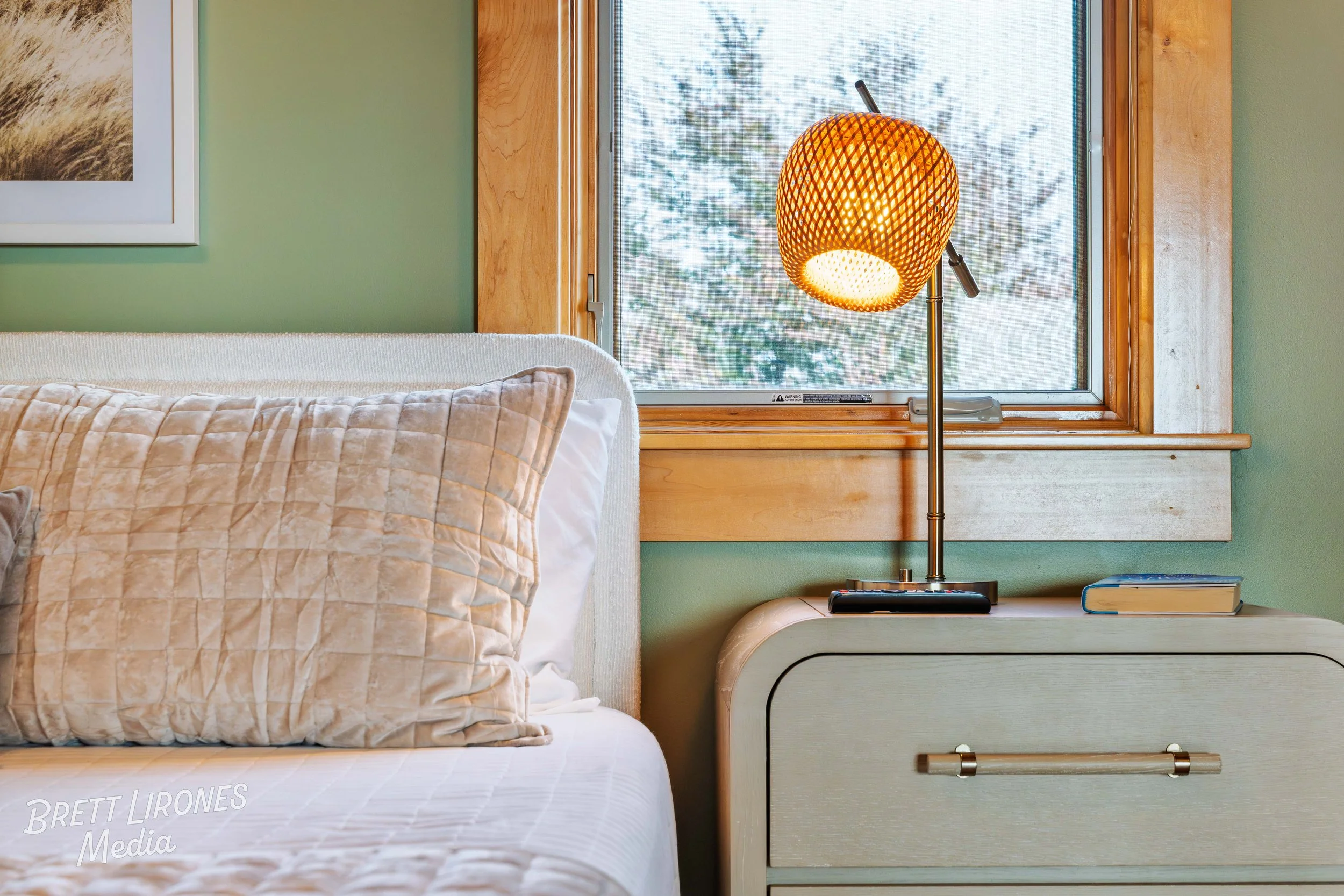 Close-up of a bed with beige quilted pillows, nightstand with a lamp, a book, and a window with a wooden frame showing trees outside.