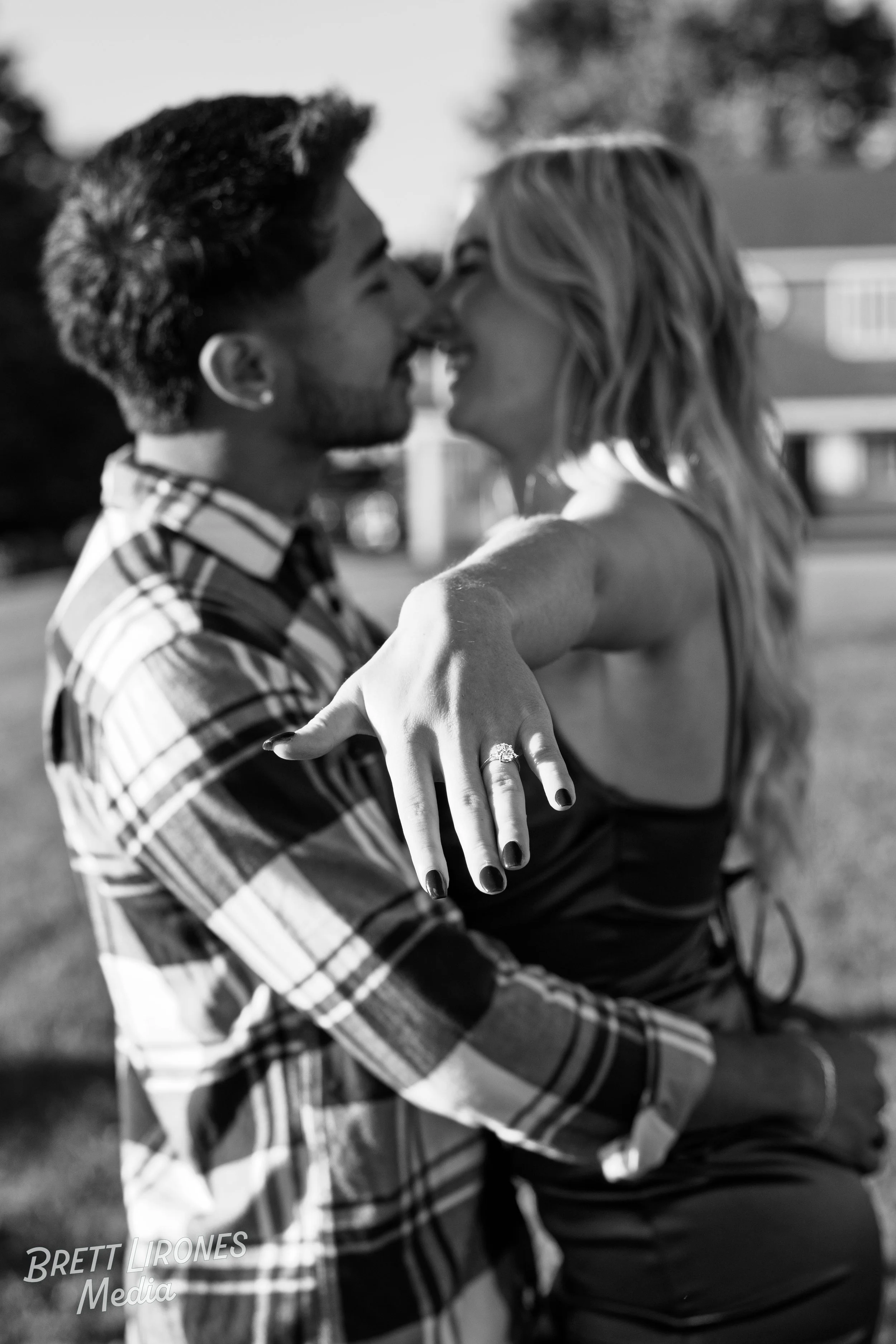 A couple sharing a kiss outdoors, with the woman showing off her engagement ring on her hand.