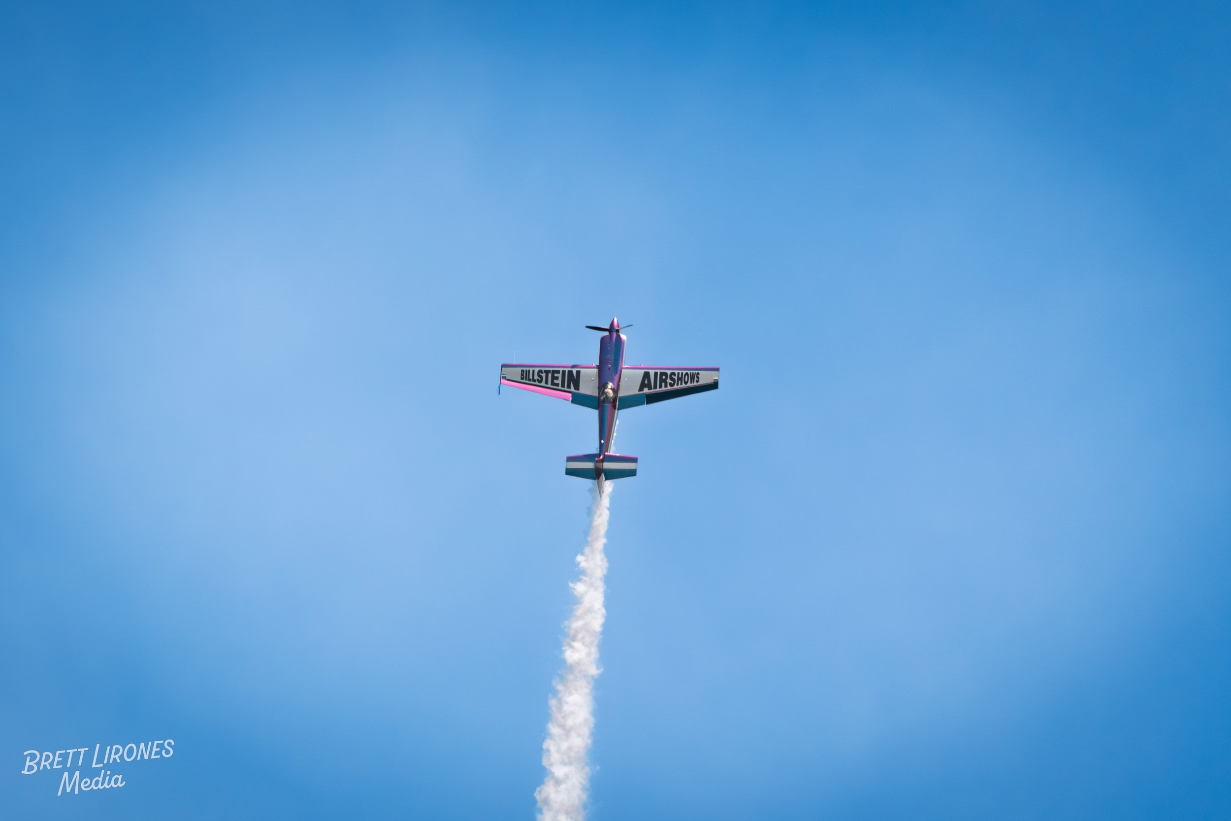 Small aerobatic airplane flying straight up with a trail of white smoke behind it against a clear blue sky.