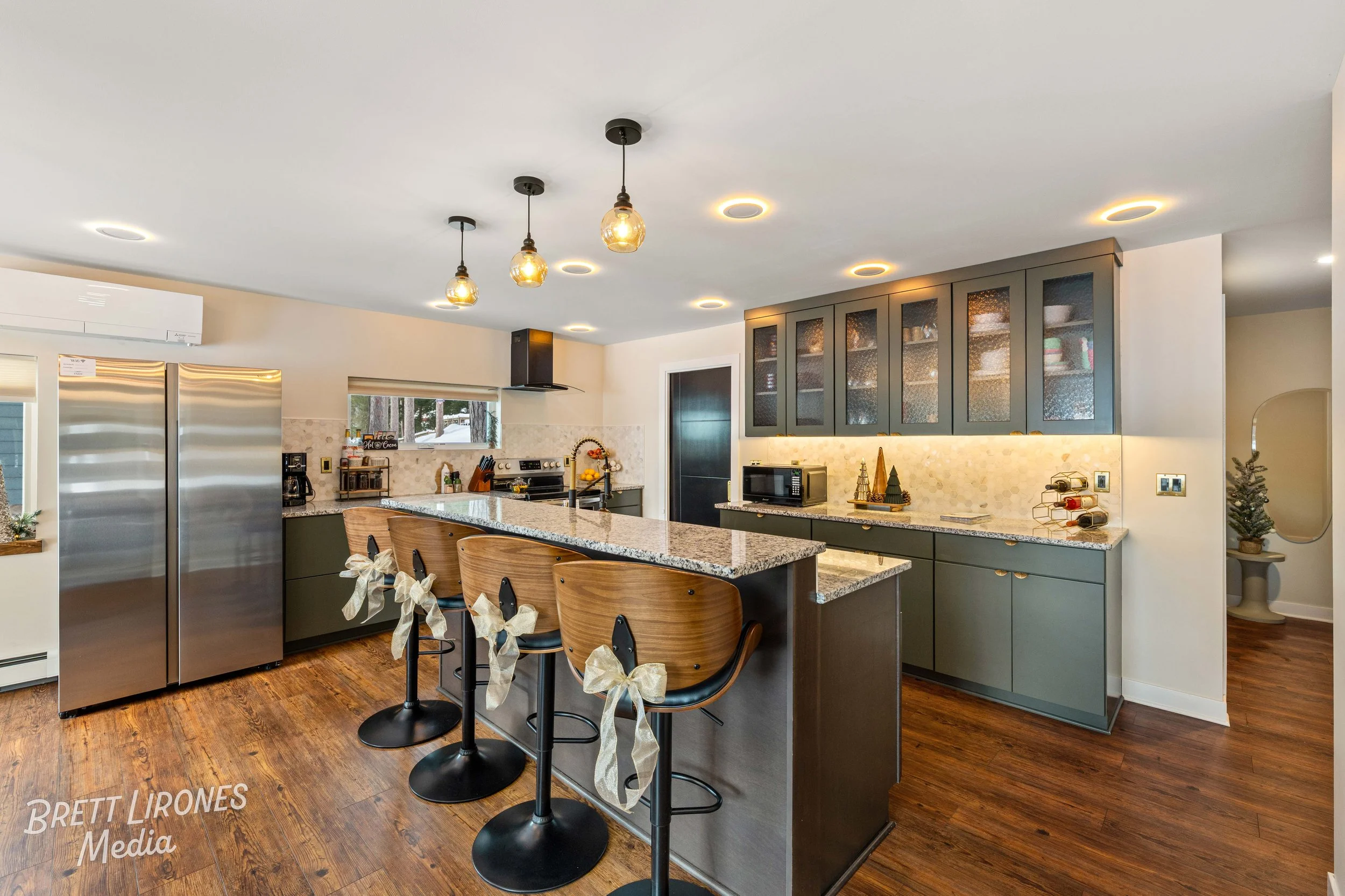 Modern kitchen with dark green cabinets, granite countertops, a central island with bar stools decorated with ribbons, stainless steel refrigerator, small window above the stove, hanging pendant lights, and Christmas decorations.