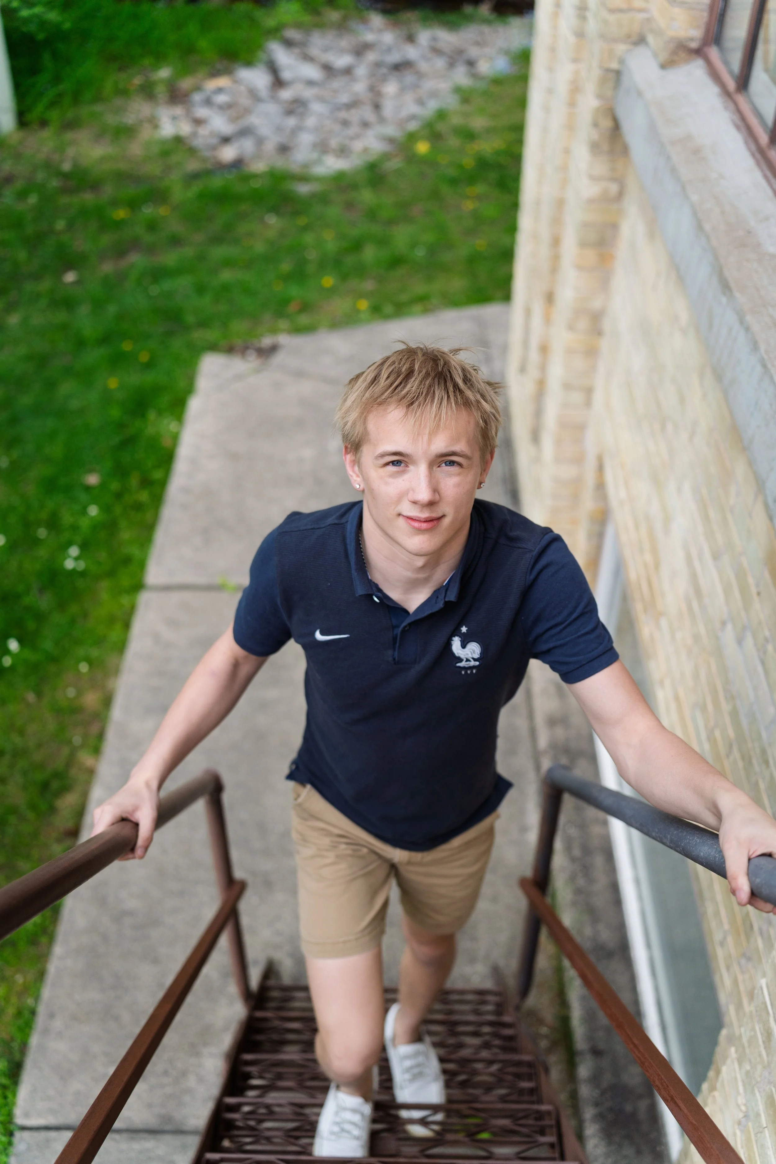 A young man with blonde hair and blue eyes standing on an outdoor metal staircase, looking up at the camera, wearing a navy blue polo shirt with a French football federation logo, khaki shorts, and white sneakers, with a background of green grass and