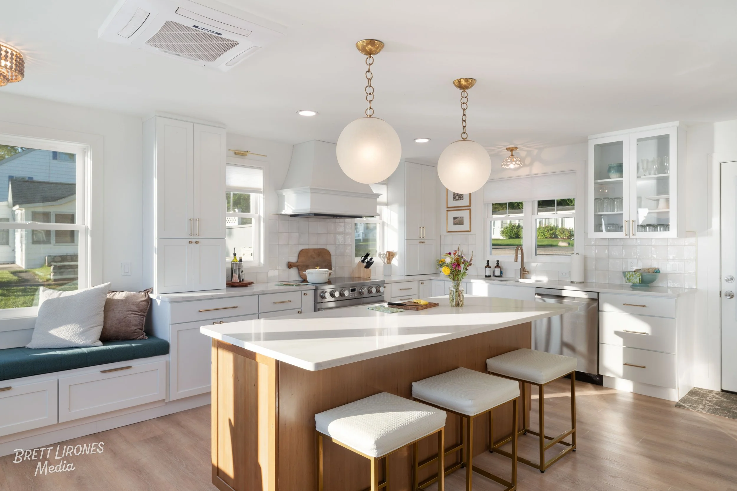 Bright, modern kitchen with white cabinetry, marble island, and large windows. There are three spherical pendant lights hanging above the island, a window seat with pillows, and open shelving with glassware. The kitchen has wood flooring and a vase o