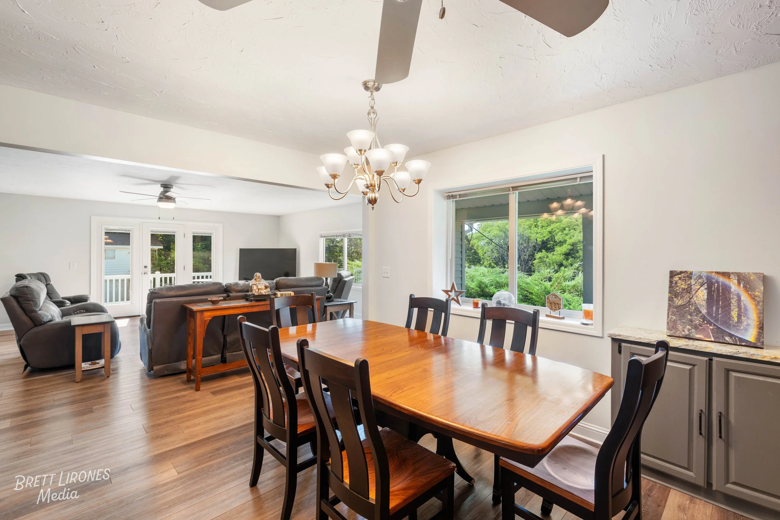 A spacious living and dining area with wooden flooring, a dark wood dining table and chairs, a gray cabinet, a large OLED TV, gray reclining sofas, and large windows showing green trees outside. A chandelier hangs above the dining table.