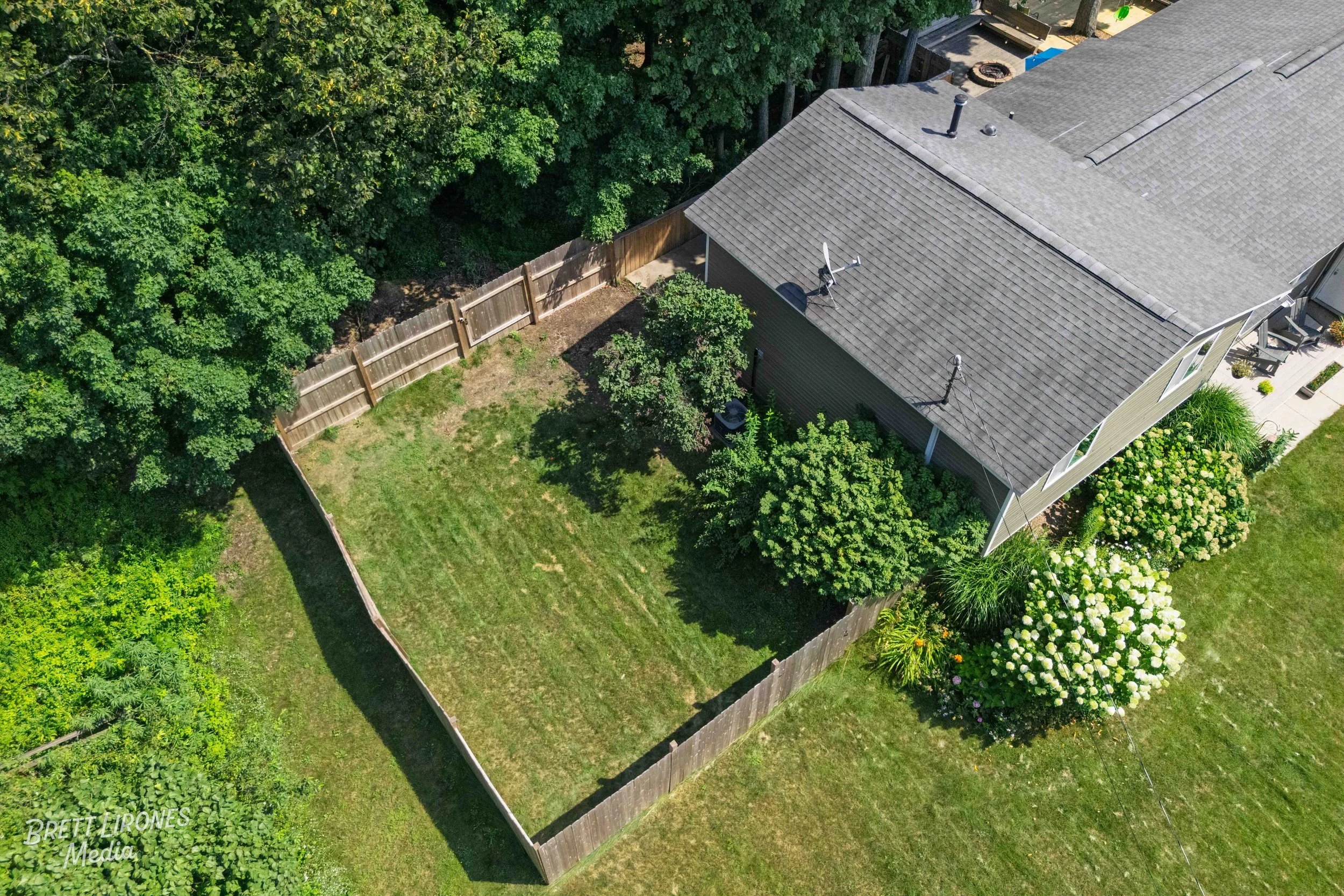Aerial view of a backyard with a wooden fence, green grass, and trees surrounding it, adjacent to a house with a gray shingled roof and a satellite dish.