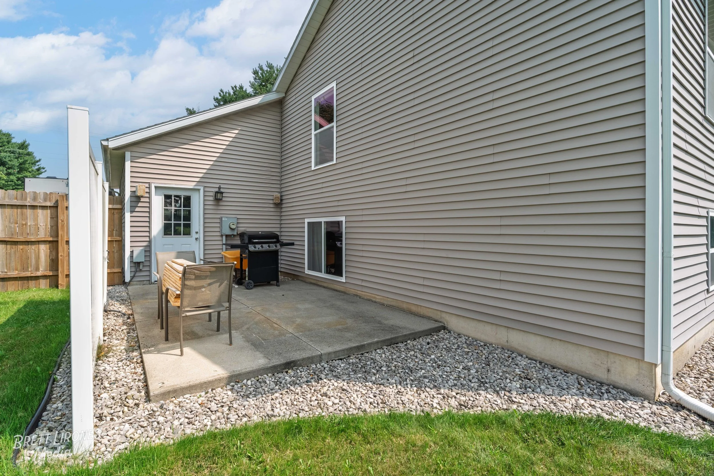 Backyard patio area with concrete slab, patio furniture, grill, and house exterior with siding, windows, and door.