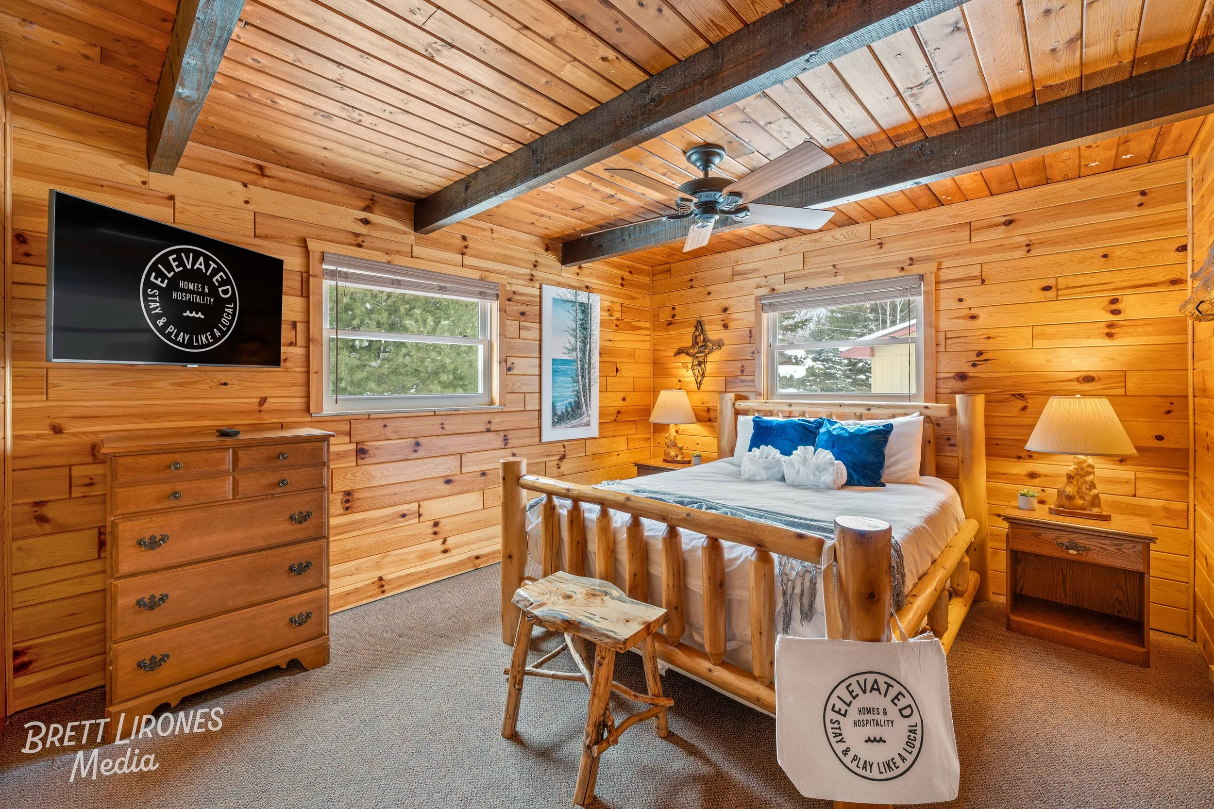 A rustic bedroom with wooden walls and ceiling, a log bed with blue pillows, two nightstands with lamps, a wooden dresser, two windows, and a ceiling fan.