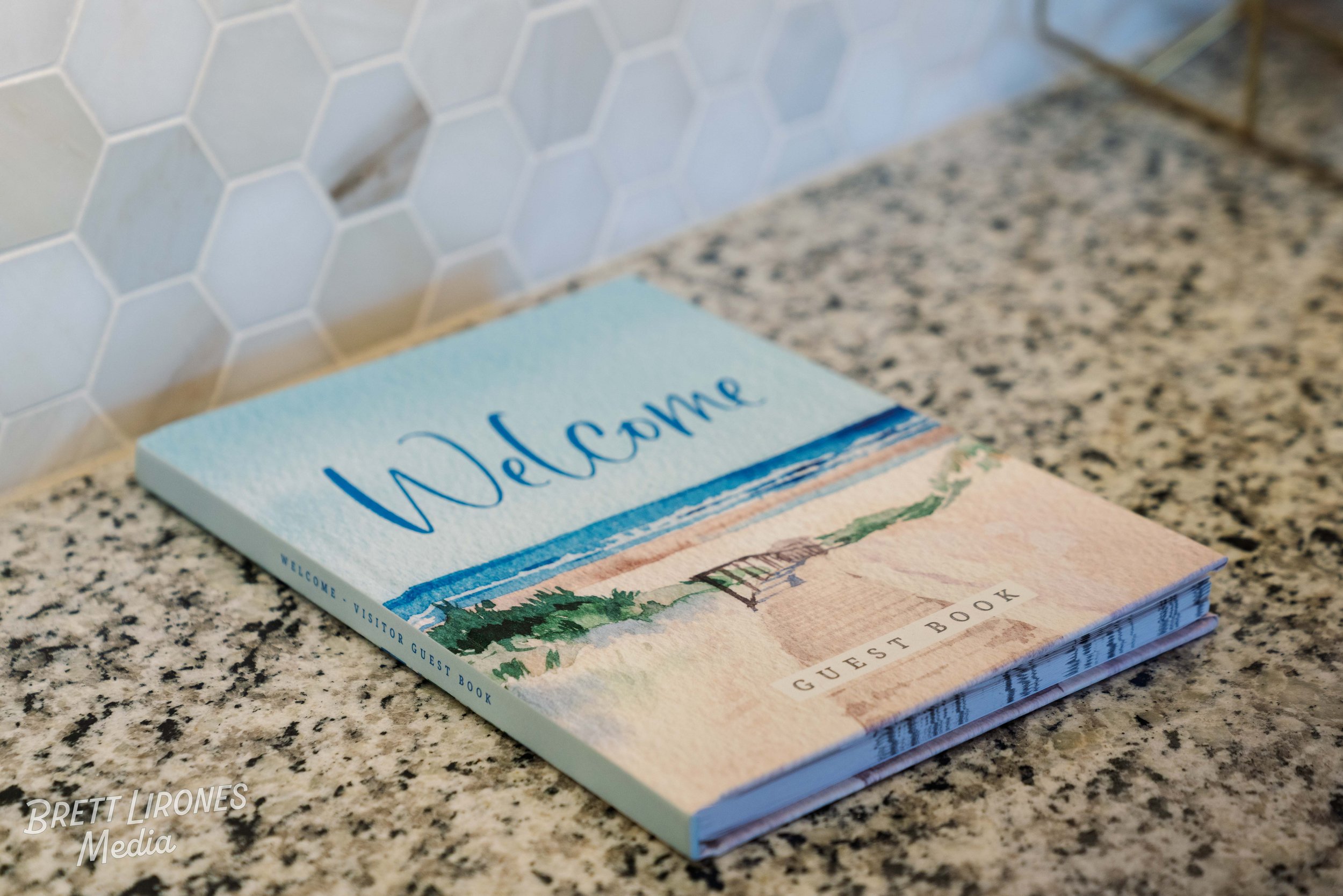 A welcome guest book with a watercolor illustration of a beach scene on the cover, placed on a speckled granite countertop with white hexagonal tiled wall in the background.