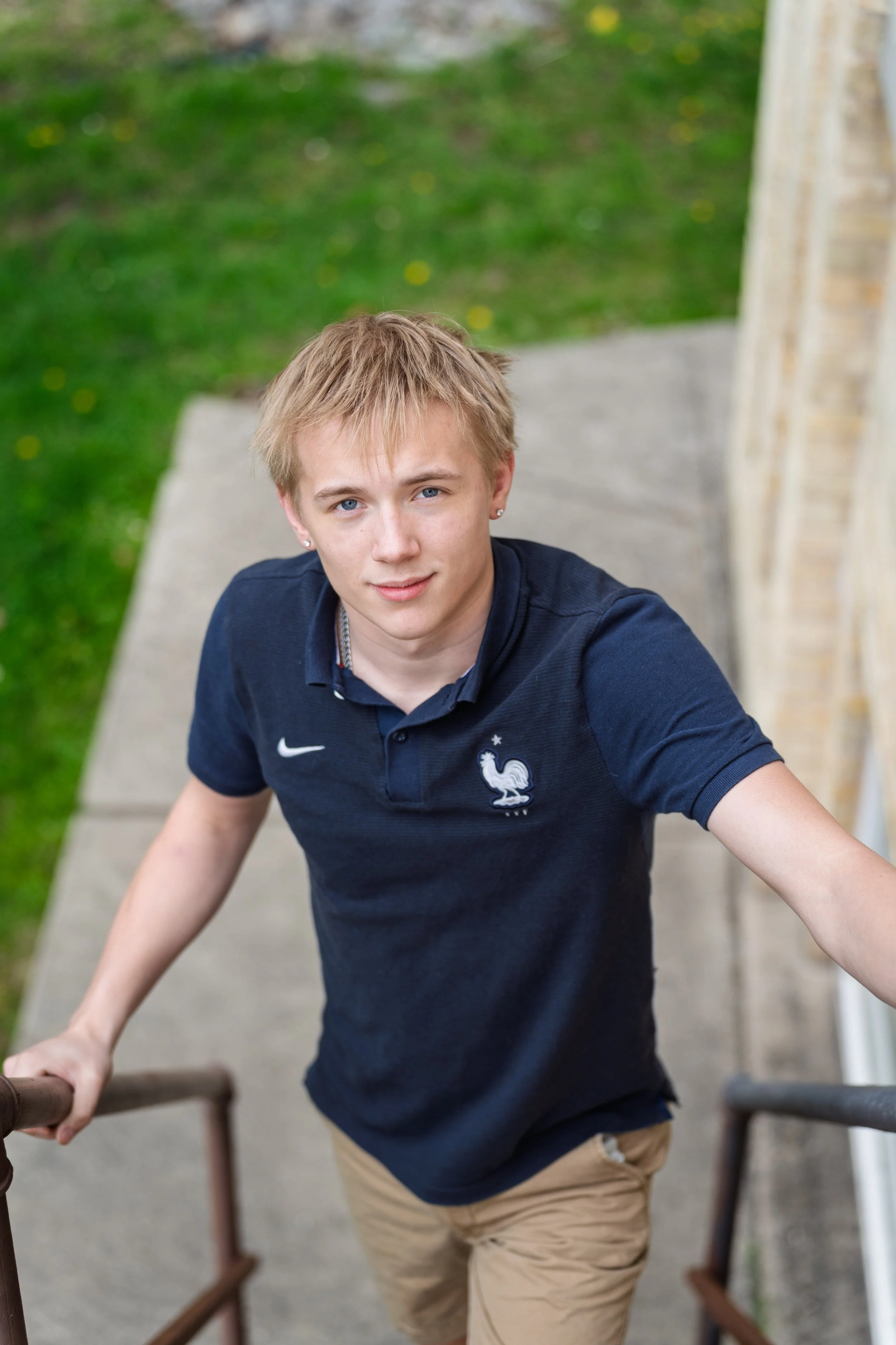 A young man with blond hair and fair skin leaning on a railing, looking up at the camera with a slight smile, wearing a navy blue polo shirt with a rooster emblem and beige pants, standing outside near a brick building with a grassy area in the backg