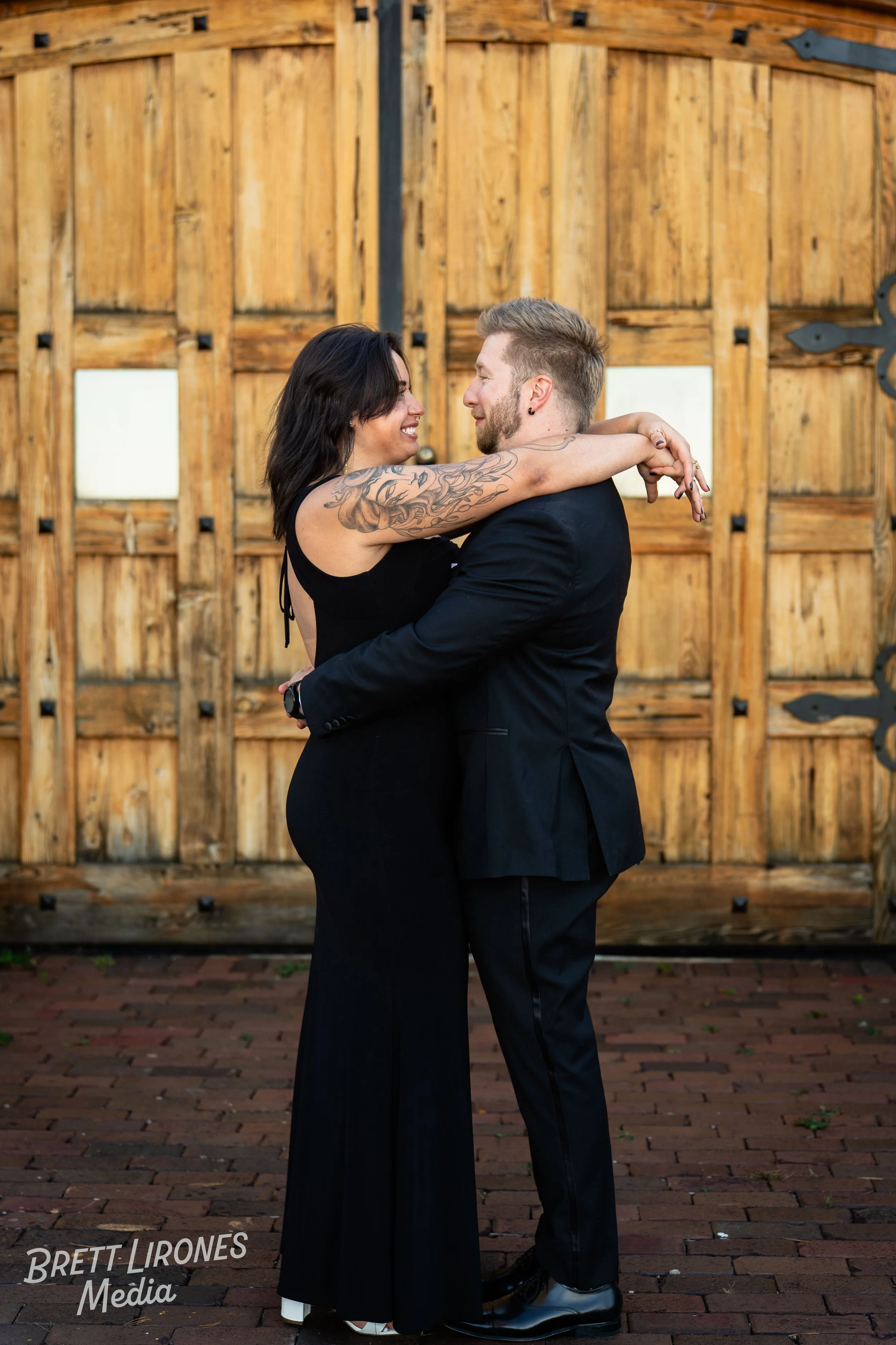 A couple dressed in black embrace and smile at each other in front of a wooden gate, with the woman’s arms wrapped around the man’s neck.