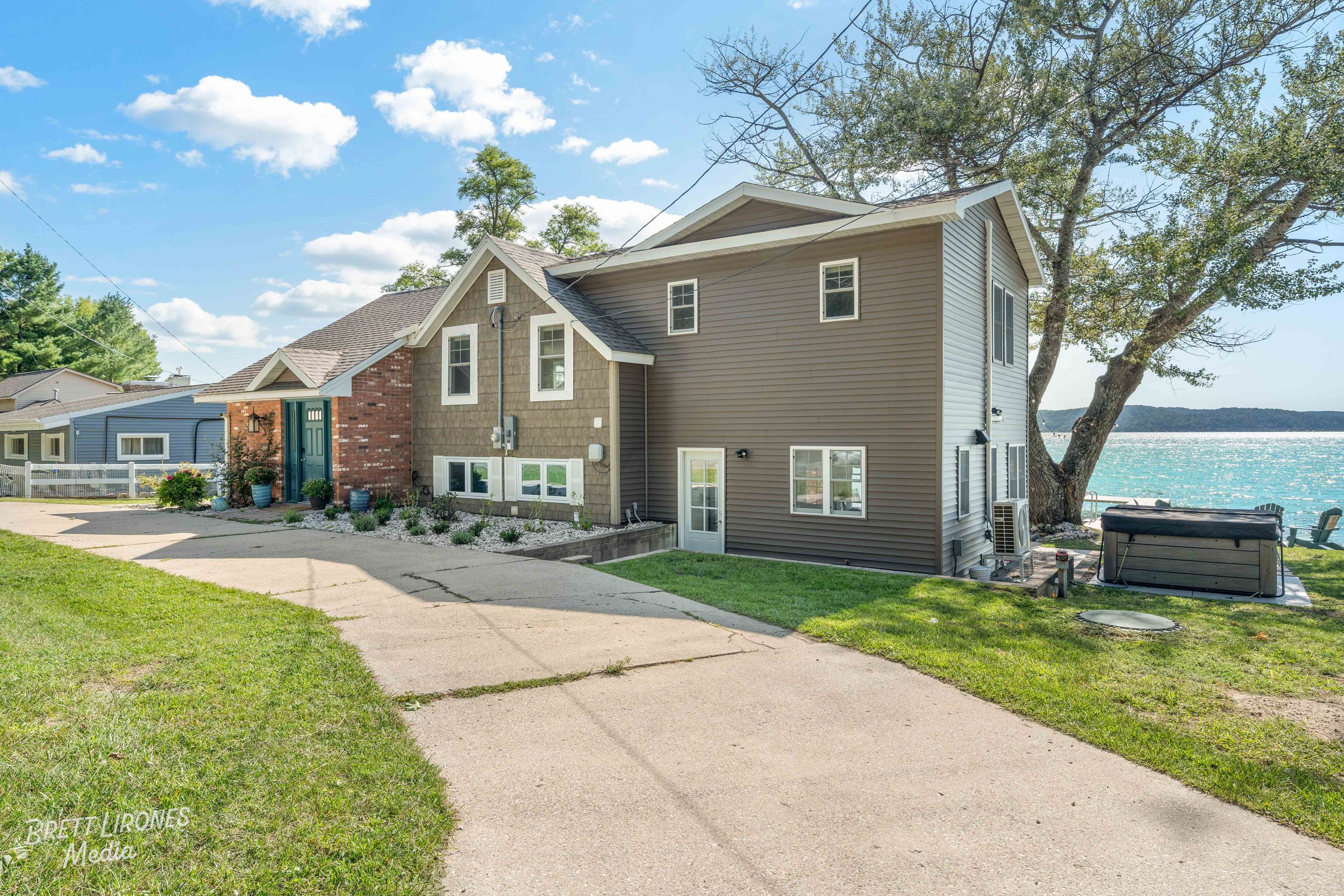 A house next to a body of water with a large tree, hot tub, and Adirondack chairs on the lawn.