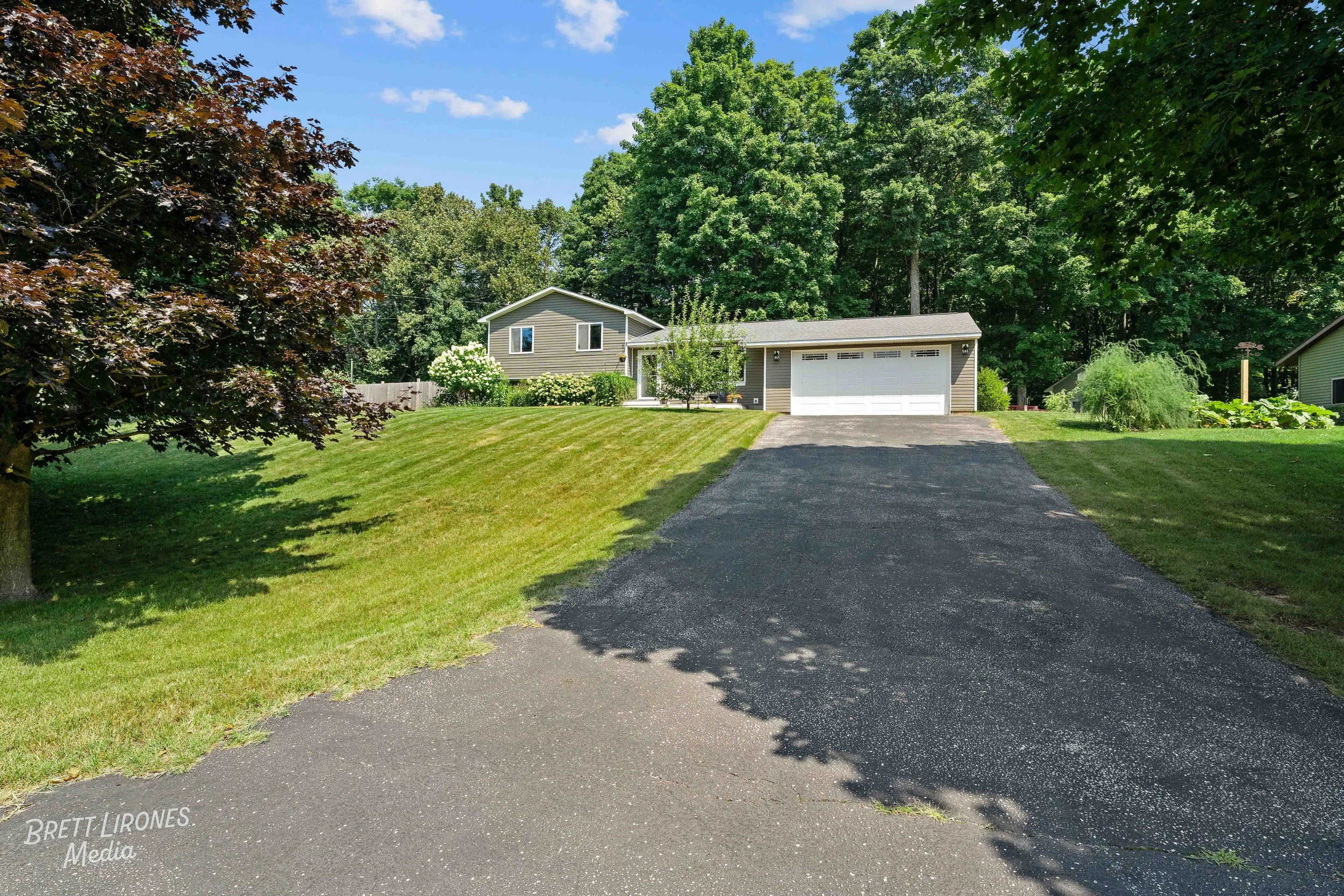 Residential house with attached two-car garage, paved driveway, well-maintained lawn, mature trees, and shrubs, under a clear blue sky.